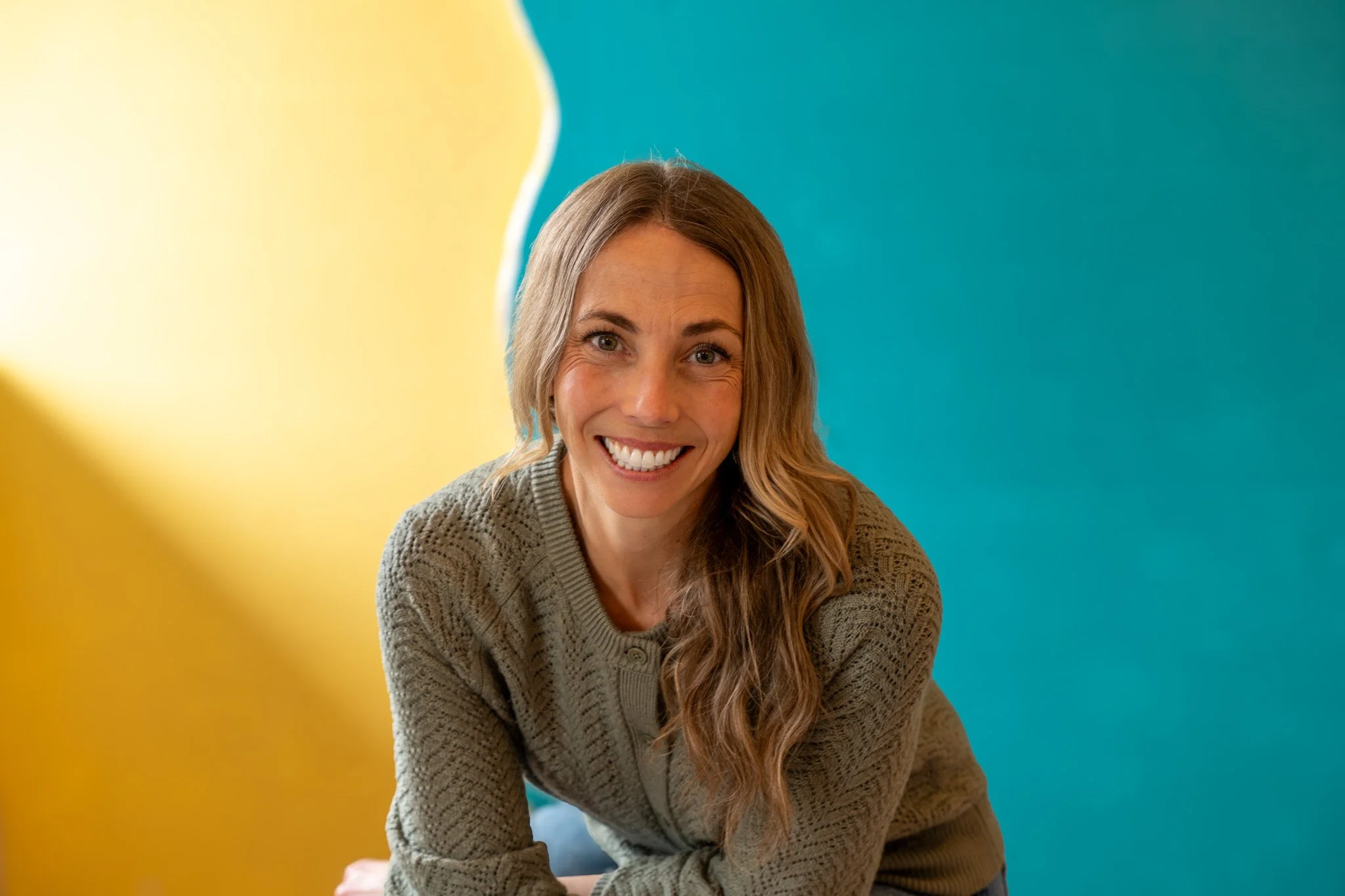 A woman smiling outdoors on a forest trail, wearing a gray beanie and a black jacket, with green moss and snow on the ground in the background.