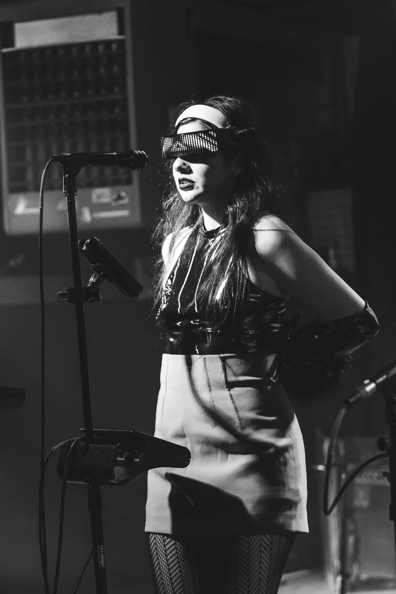 A woman with long hair wearing a headband and patterned sunglasses stands at a microphone on stage, dressed in a shiny top, skirt, fishnet tights, and long gloves in a black-and-white photograph.
