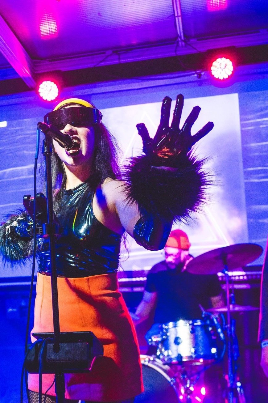 A female singer wearing futuristic sunglasses, a shiny black top, and a red skirt, performing on stage with a microphone. She has black furry gloves and is making a dynamic gesture with her hand. A drummer wearing glasses and a red cap is visible in the background, playing drums under colorful stage lights.