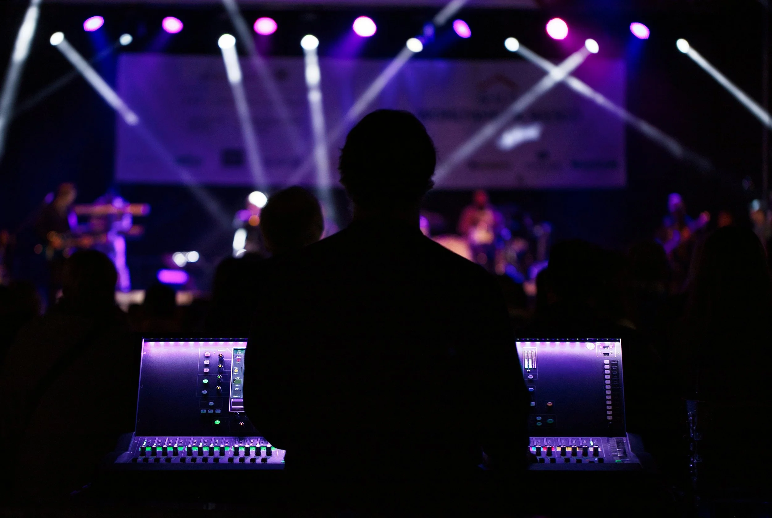 A concert scene with a sound technician working at a mixing console in the foreground, and a band performing on stage with purple lighting and spotlights in the background.