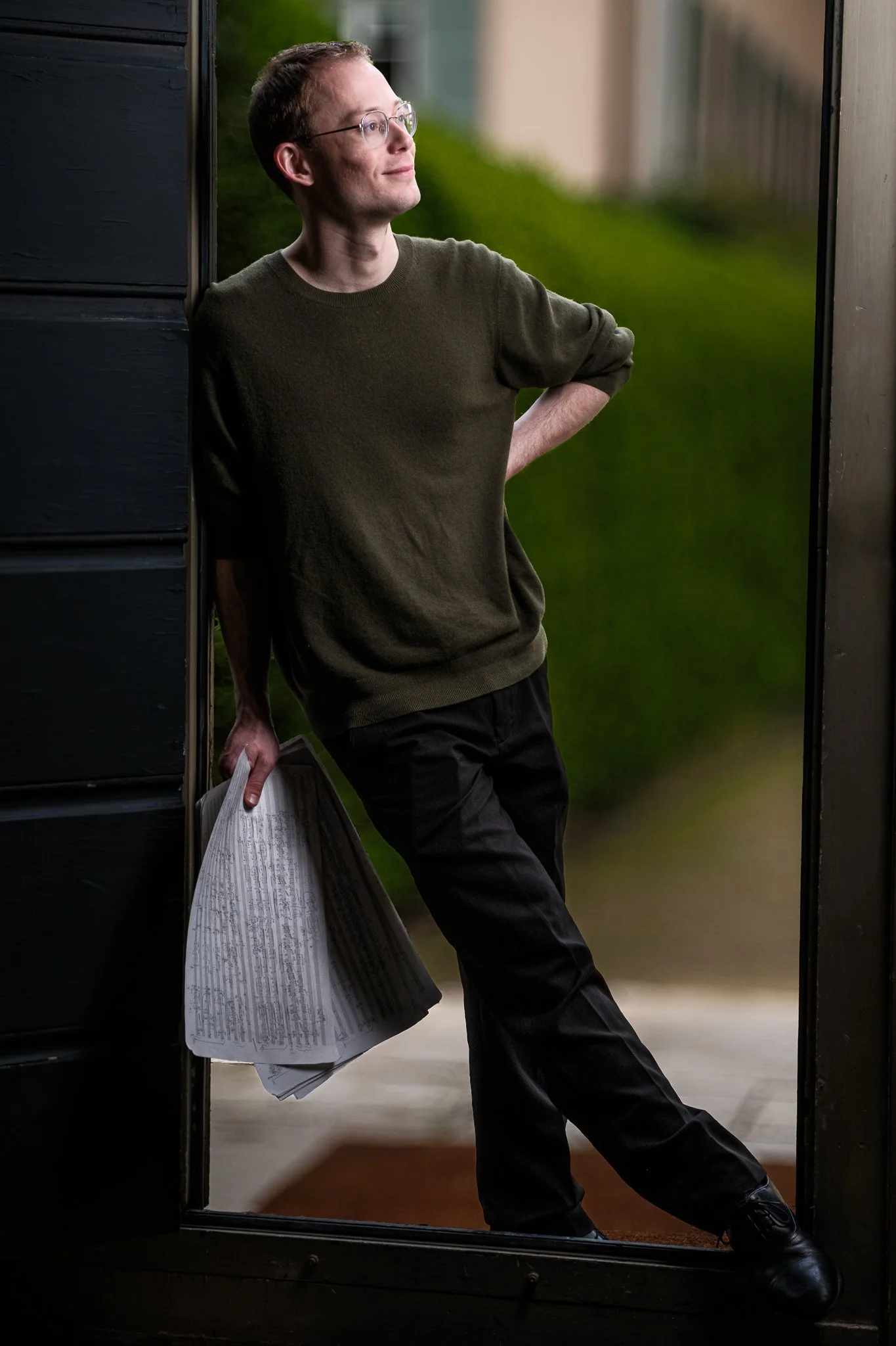Composer Miles Walter stands in a doorway, looking away, looseleaf handwritten manuscript score of his Piano Quartet in hand.