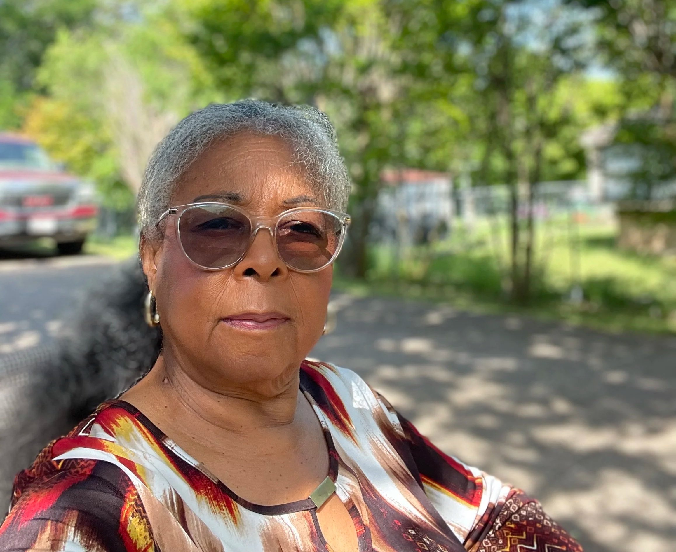 An older Black woman with gray hair wearing sunglasses and a colorful patterned top, standing outdoors with trees, houses, and a parked car in the background.