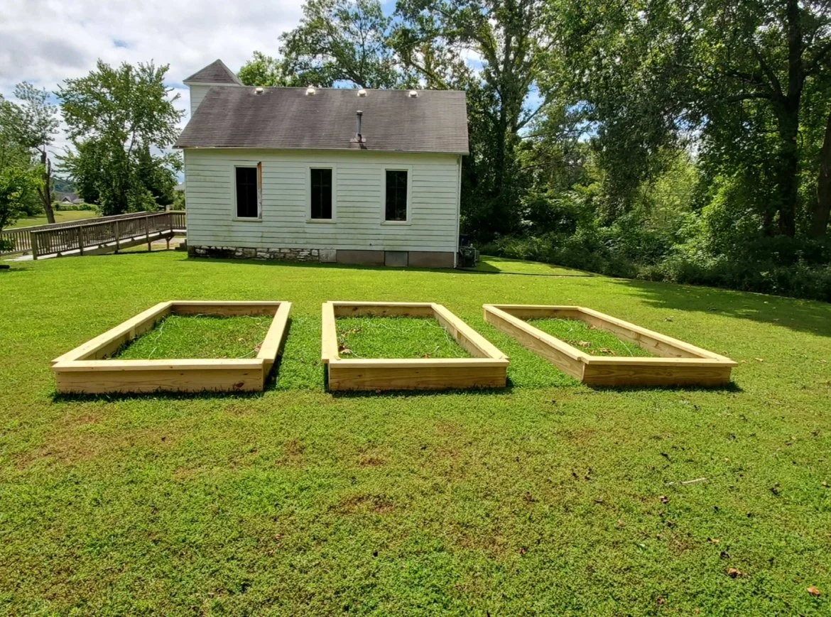 Three empty wooden raised garden beds on a grassy yard in front of a white house surrounded by trees.