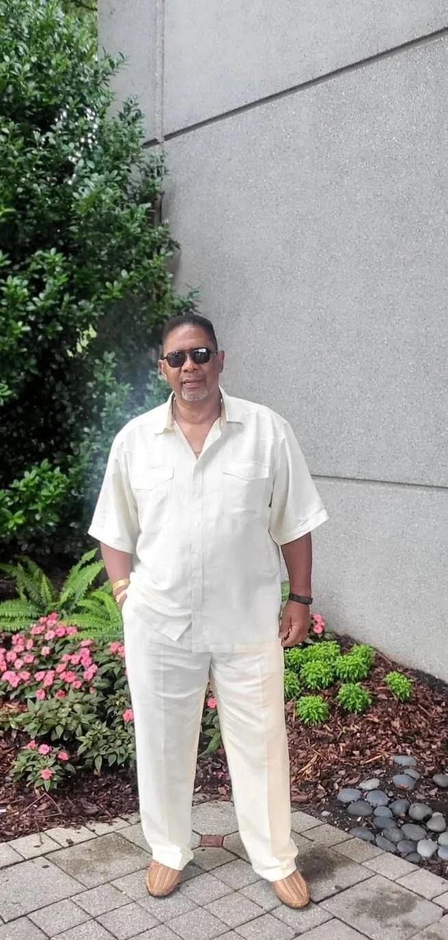 A Black man stands outdoors near a gray concrete wall and green shrubbery with pink flowers, wearing a white short sleeve shirt, white pants, tan shoes, black sunglasses, and a black wristwatch.