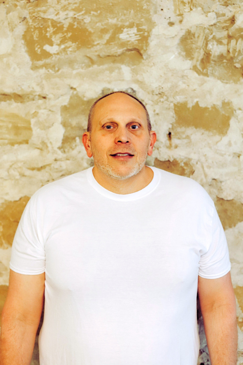A smiling man, wearing a white t-shirt, standing in front of a rustic stone wall.