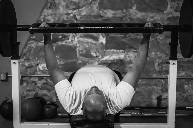 A man lifting a barbell while lying on a bench press at the gym with kettlebells nearby.