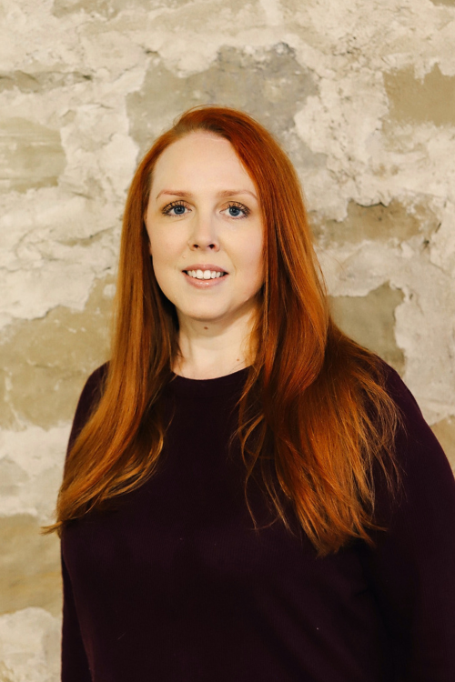 A woman with long red hair smiling at the camera, standing in front of a textured stone wall.
