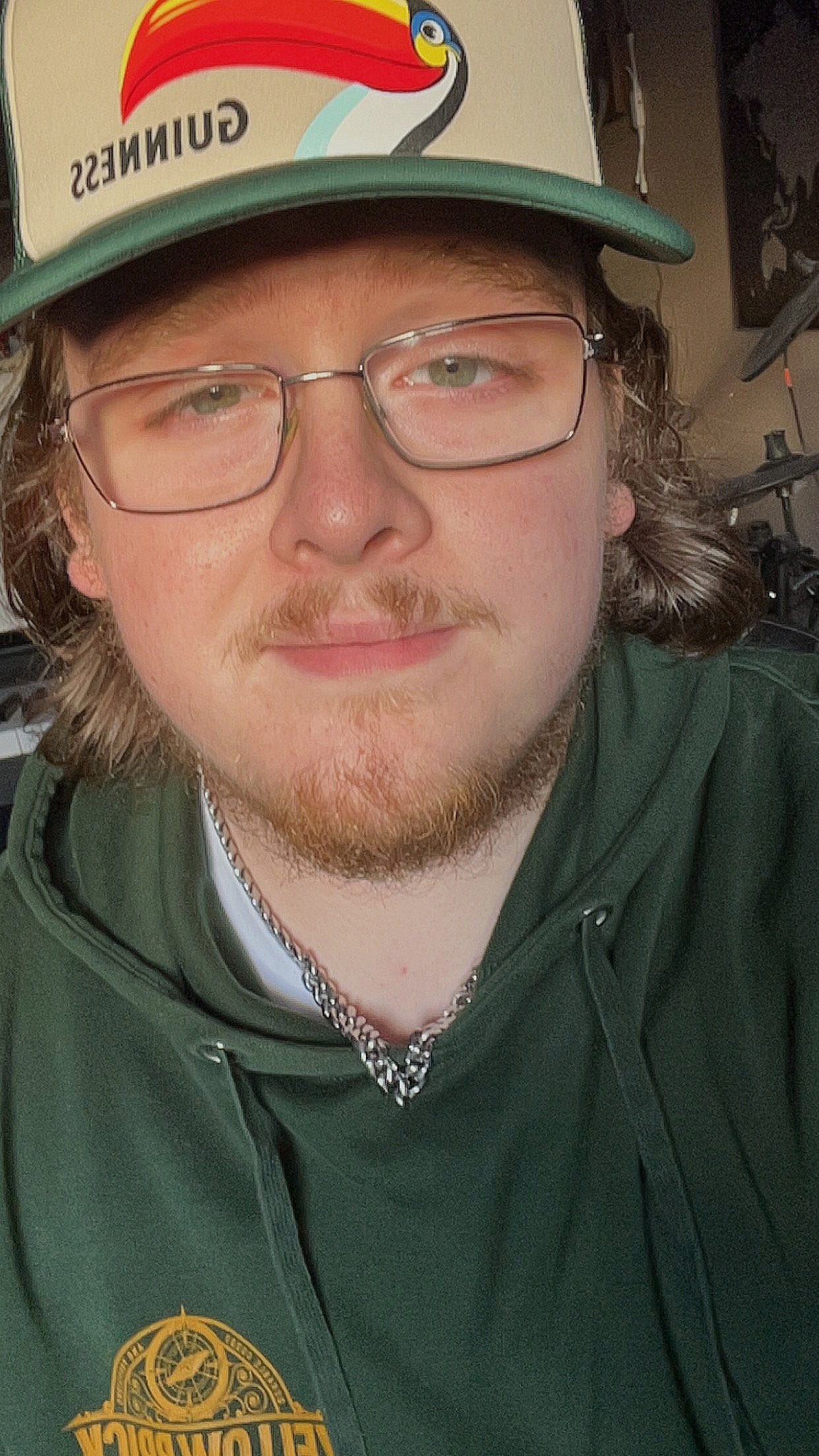 A young man with glasses, a beard, and long hair wearing a green hoodie with a logo, sitting indoors, wearing a hat with a toucan and the word 'GUNNES'S' on the logo.