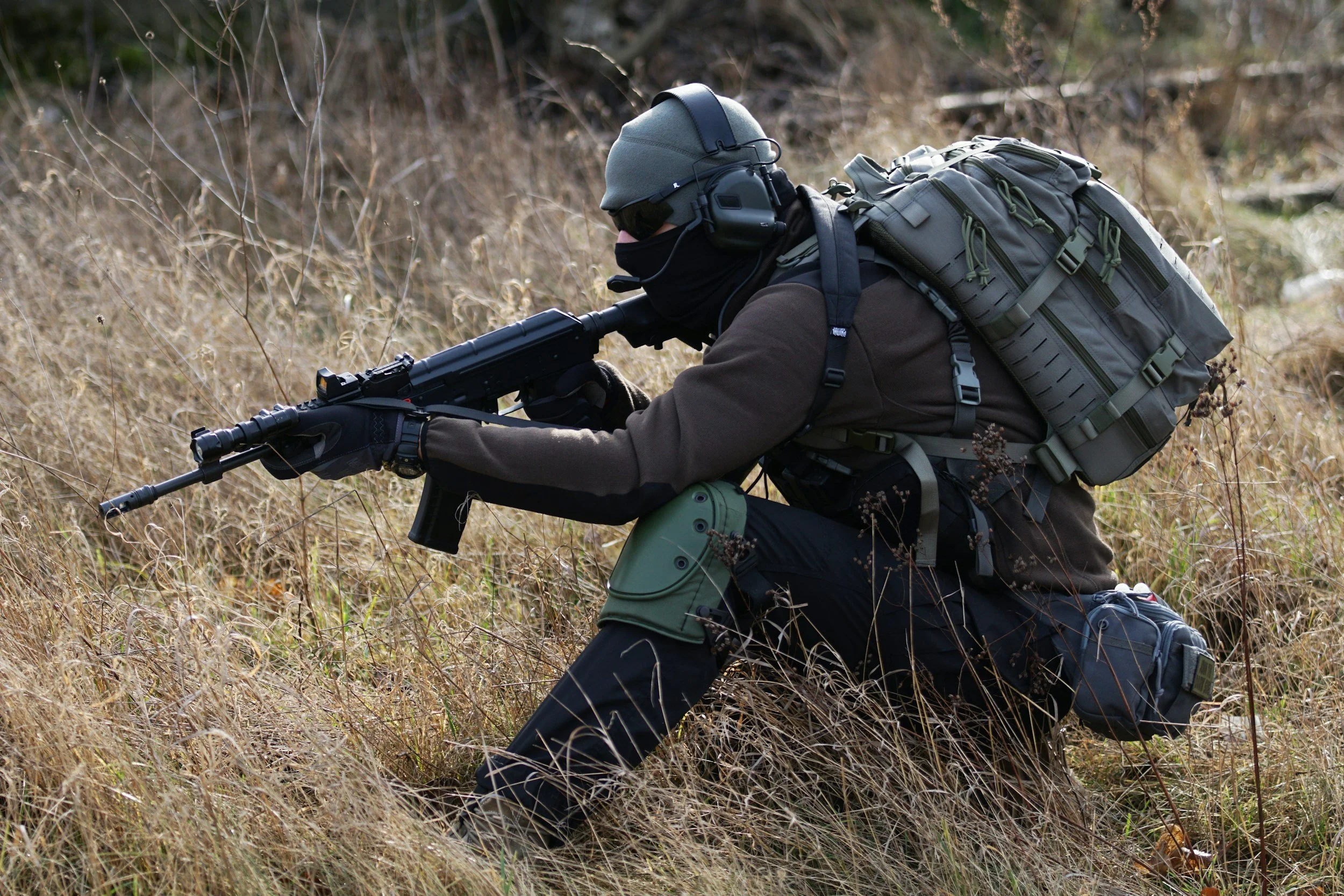 A person dressed in tactical gear, kneeling in tall grass, aiming a rifle in a wooded area.