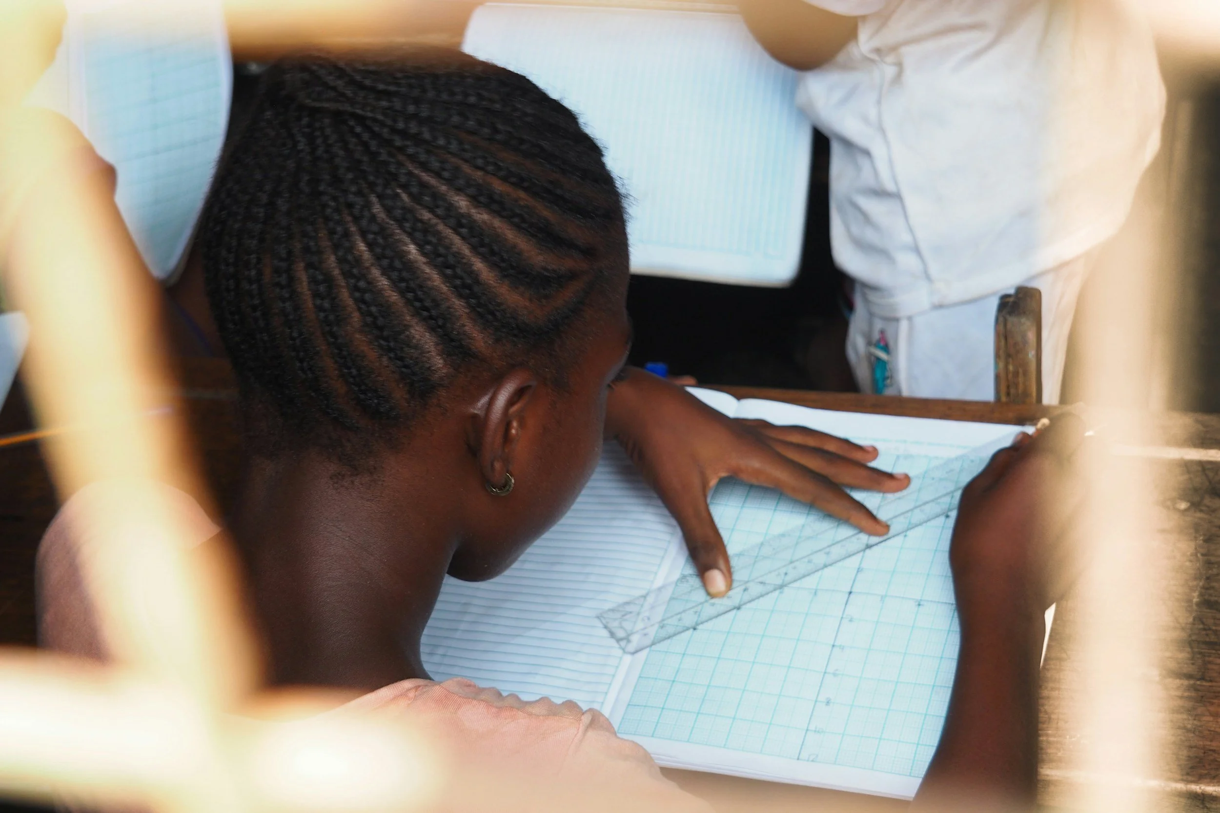 A girl with braided hair working on a grid-lined notebook with a ruler at a desk.