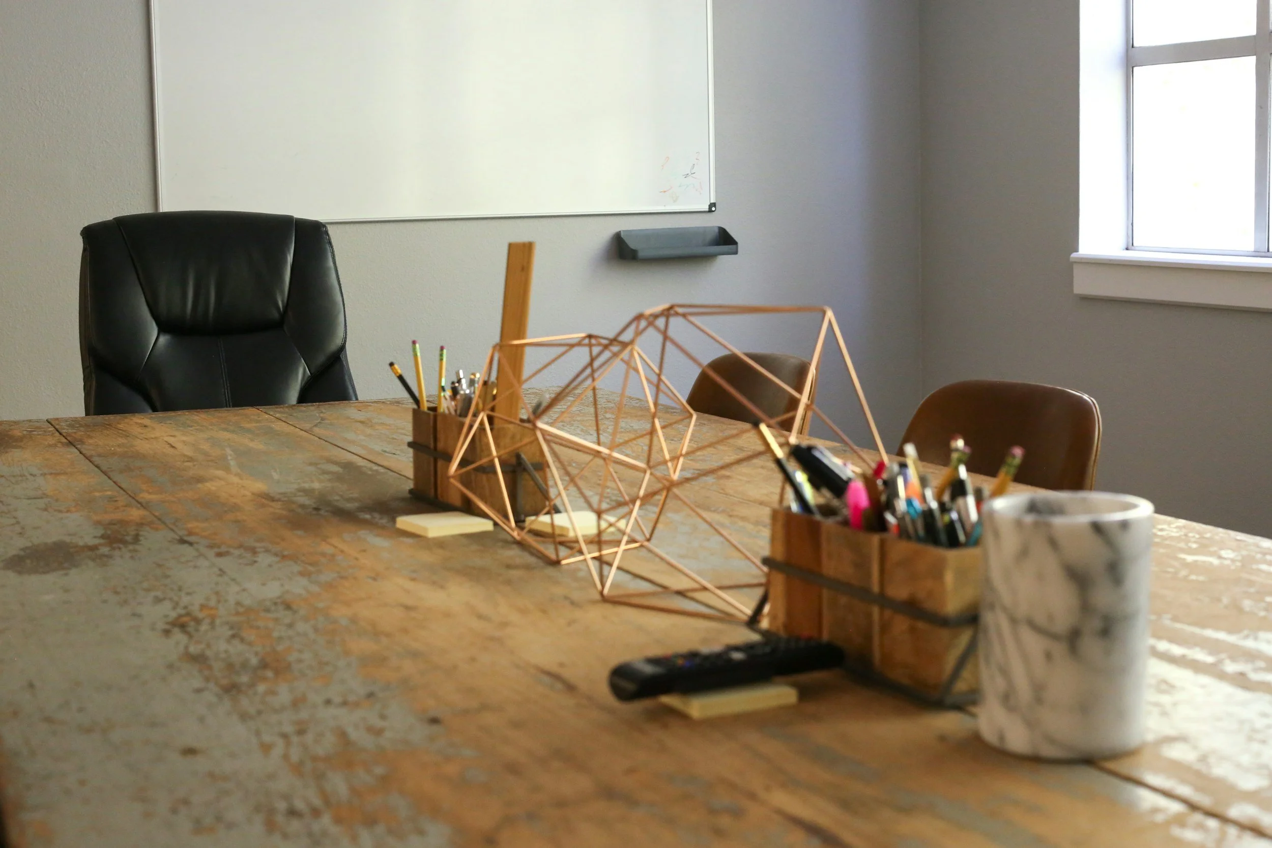An empty conference room table with a black leather chair, a computer monitor, and various office supplies including pencils, pens, markers, sticky notes, a remote, and a marble-patterned mug, with a window and a whiteboard on the wall.