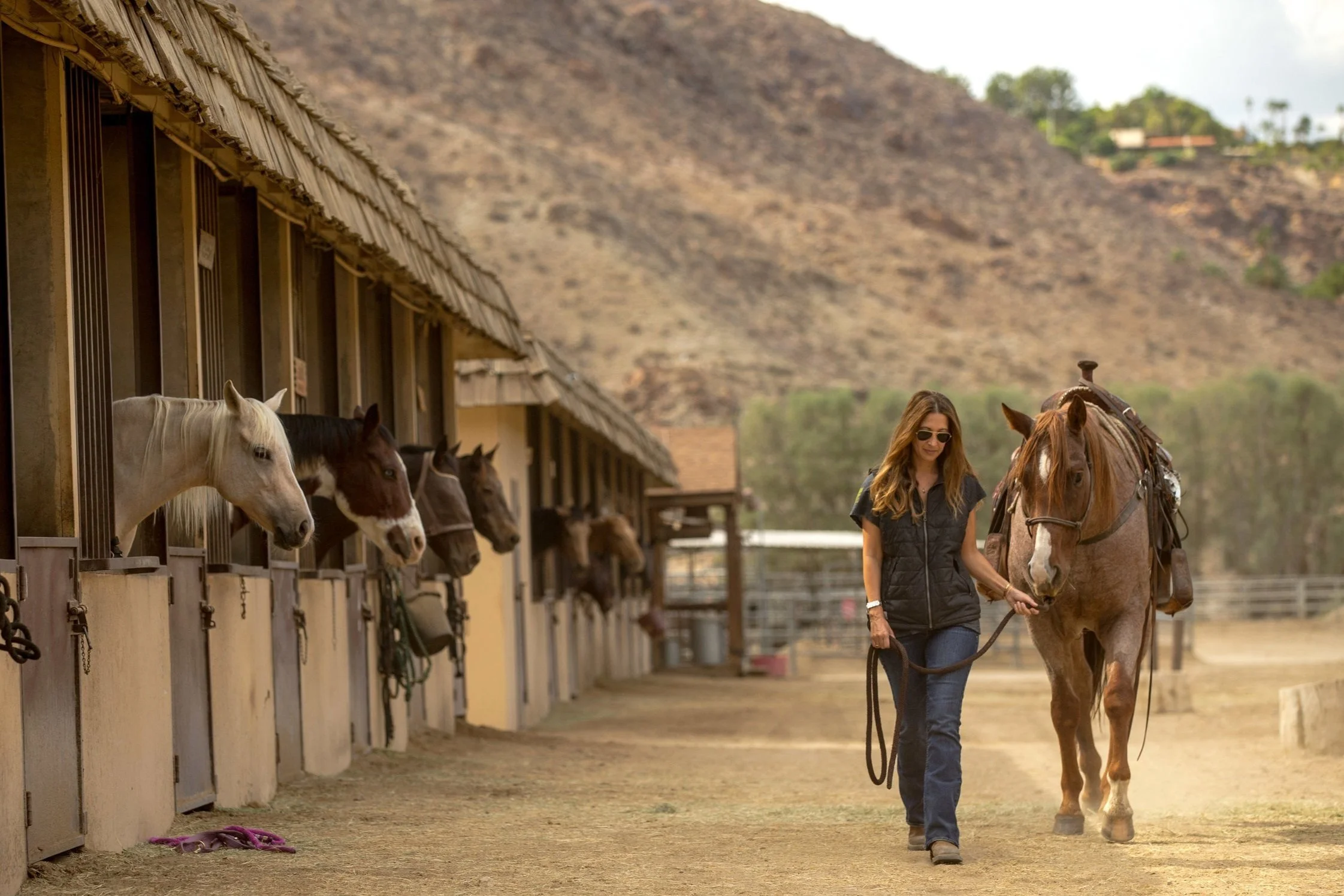 A woman with long hair, sunglasses, and casual clothing walking a horse on a dirt path next to a stable with multiple horses poking out of their stalls, set against a mountain landscape.