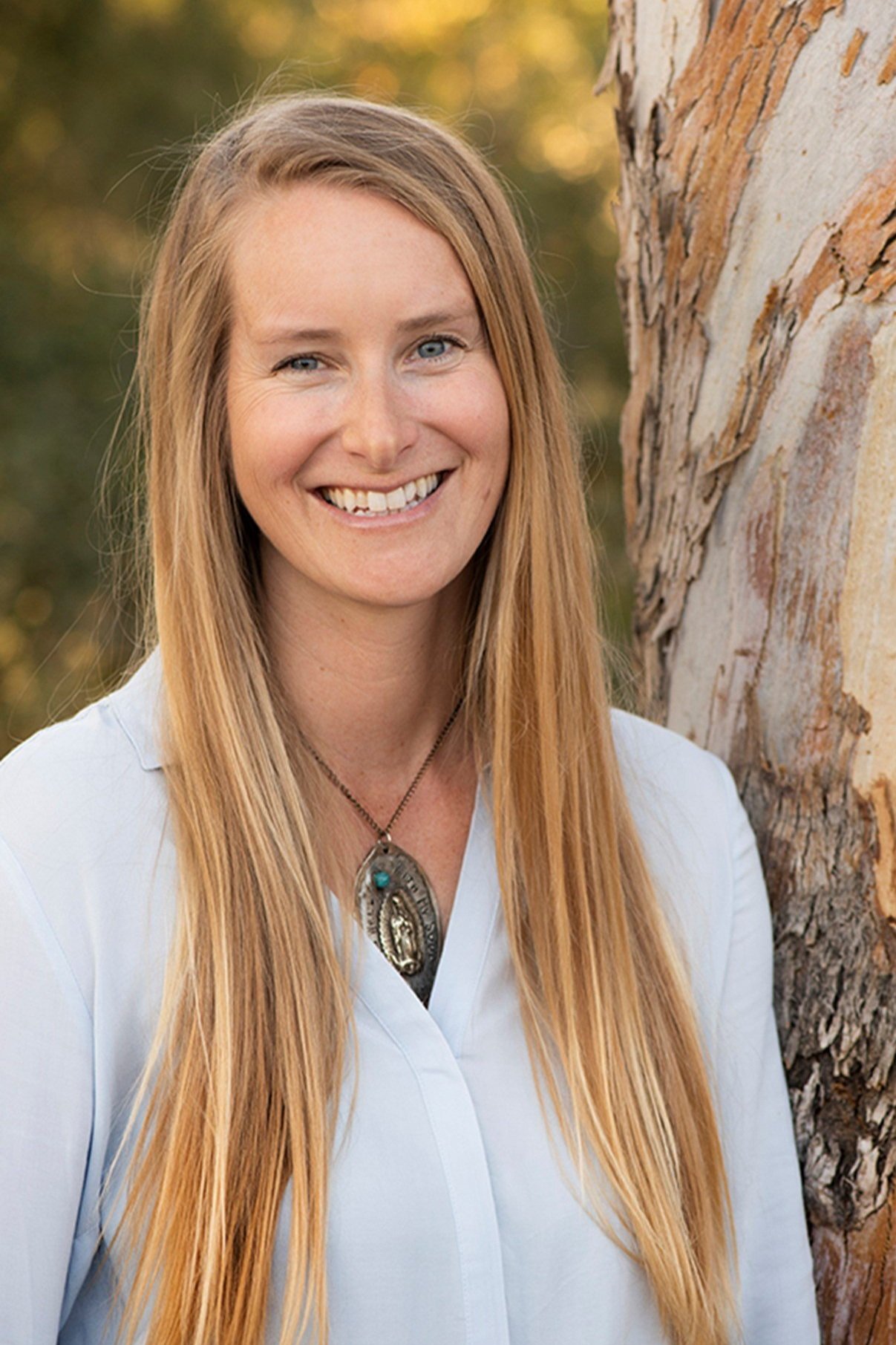 A woman with long red hair and blue eyes smiling outdoors next to a tree during sunset.