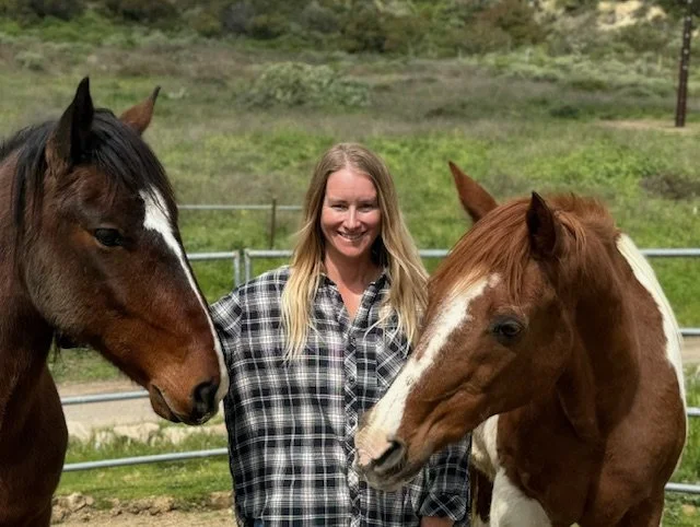 April with long blonde hair wearing a plaid shirt standing between two horses in a fenced pasture, smiling at the camera.