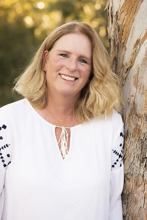 A smiling woman with blonde hair in a white blouse with black embroidery, leaning against a tree in an outdoor setting with blurred greenery in the background.