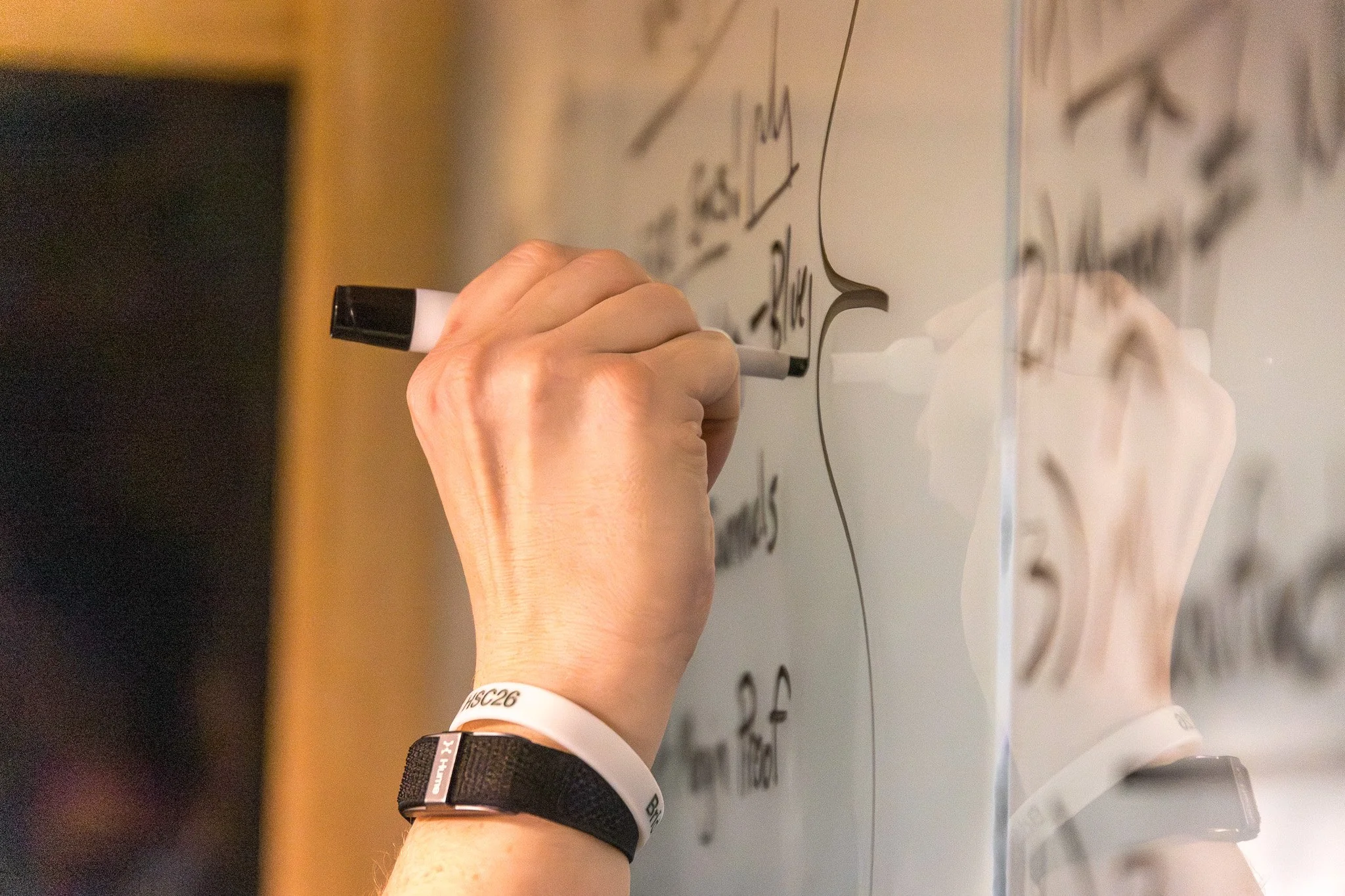 Person writing on a glass whiteboard with a black marker, with notes and diagrams, and a reflection of their hand and part of their body visible.
