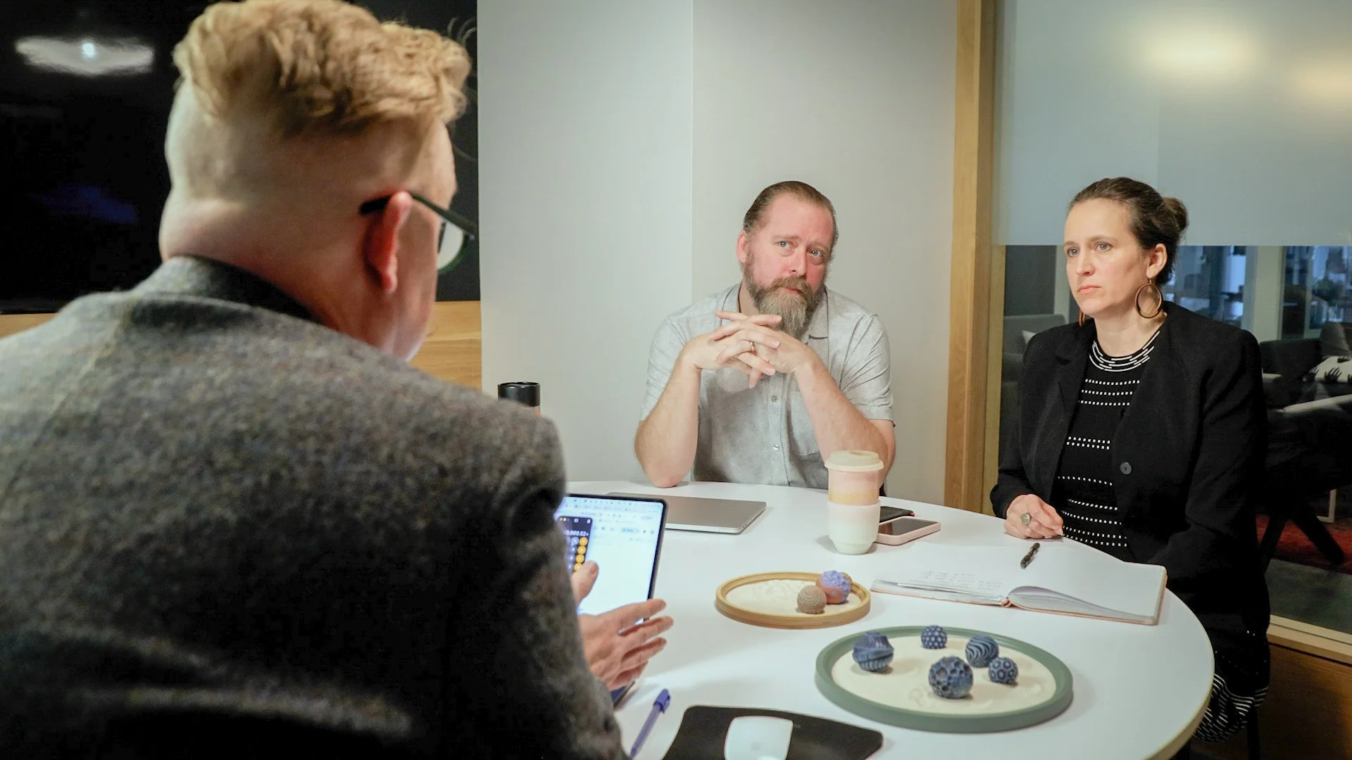 Three people sitting at a round table in a modern office or conference room, engaged in a discussion. The person on the left, with glasses and reddish hair, is facing away from the camera. The man in the middle has a beard and long hair tied back, wearing a light-colored shirt. The woman on the right has dark hair pulled back, wearing a black blazer and a striped shirt. On the table are notebooks, a coffee cup, and decorative items resembling patterned rocks or objects.
