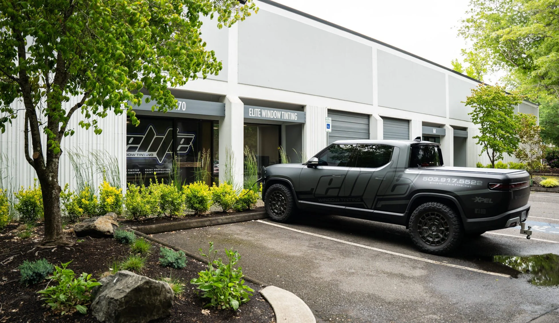Parking lot outside a building with a sign that reads 'Elite Window Tinting.' A black pickup truck is parked in a parking space near the building, with trees and shrubs in the landscaped area surrounding the building.