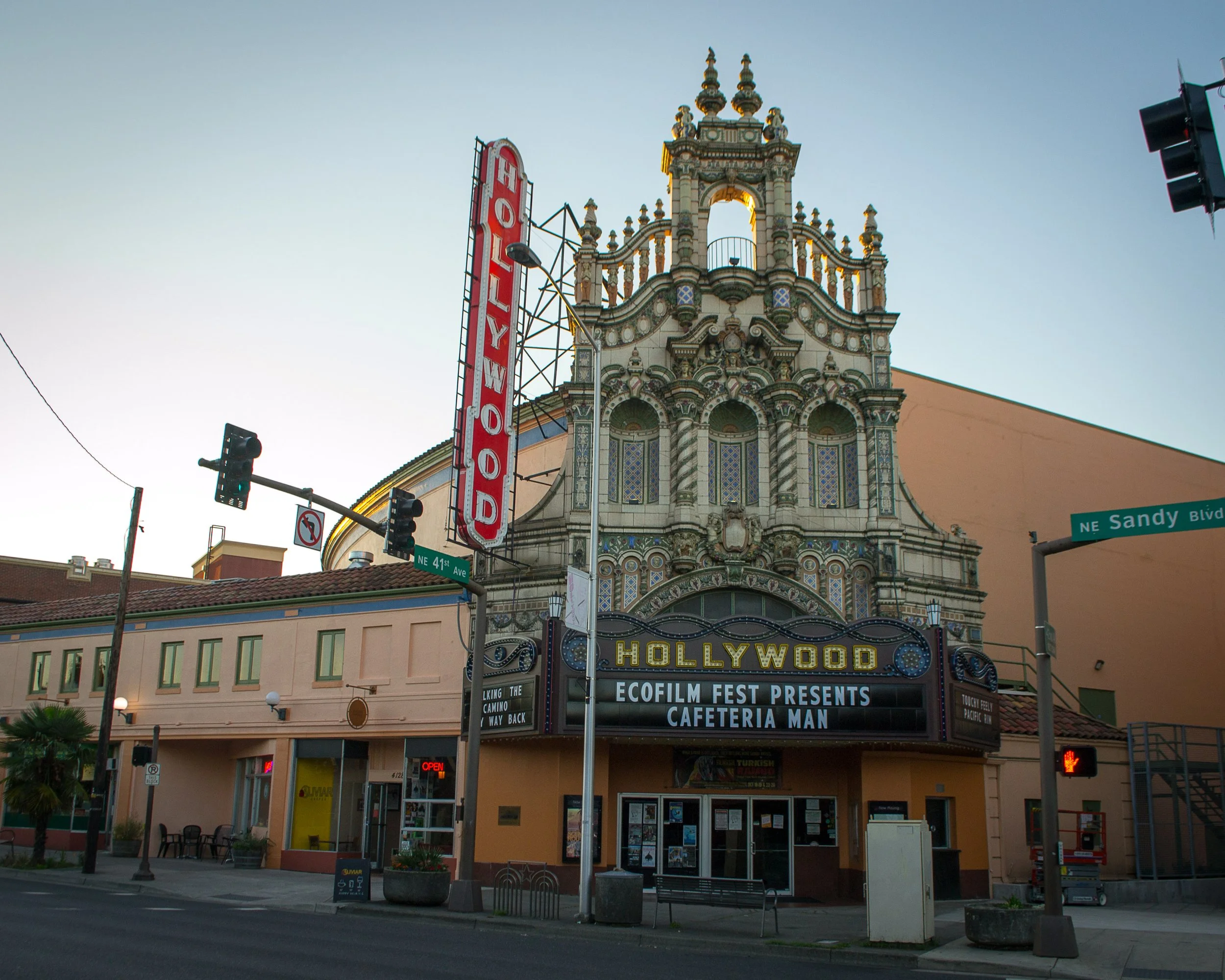 The historic Hollywood Theatre in Portland, Oregon, with a decorative facade and a marquee announcing an Ecofilm Fest event. Traffic lights and street signs are visible at the intersection of NE 41st Ave and NE Sandy Blvd.