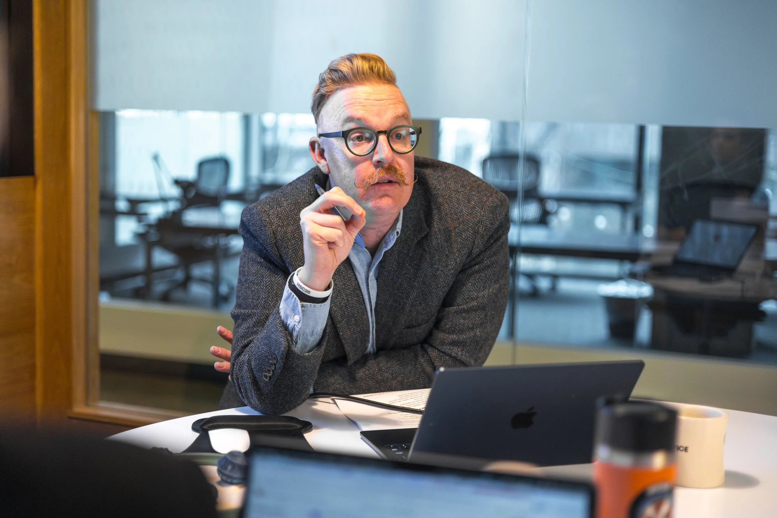 A man with glasses and a mustache, wearing a gray blazer over a light blue shirt, sitting at a conference table in front of a laptop, with a serious expression and holding a pen near his face in a modern office conference room.