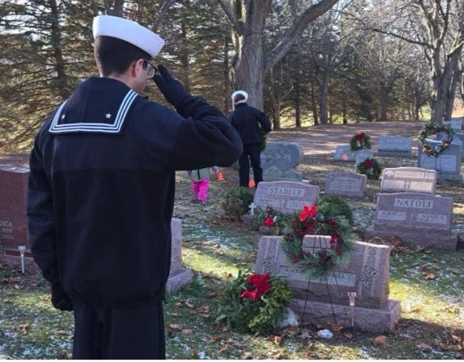 Two individuals, dressed in Navy uniforms, paying respects at a cemetery with gravestones adorned with flowers and wreaths.