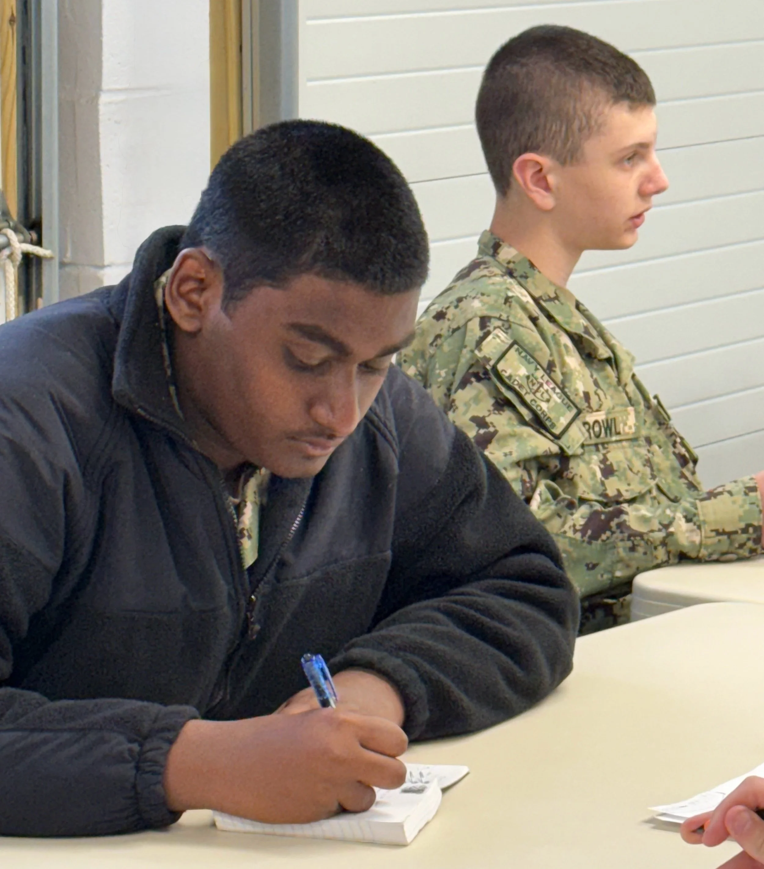 Two young men sitting at a table, one writing in a notebook, the other wearing a military uniform.