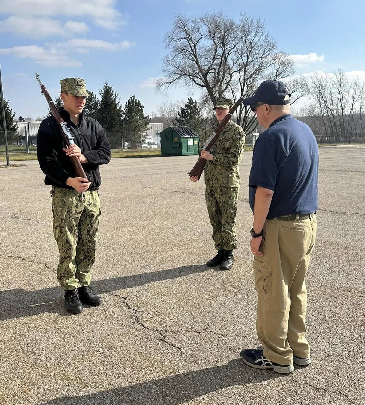 Two cadets in camouflage uniforms holding rifles stand in formation while Executive Officer instructs them in color guard riflery.