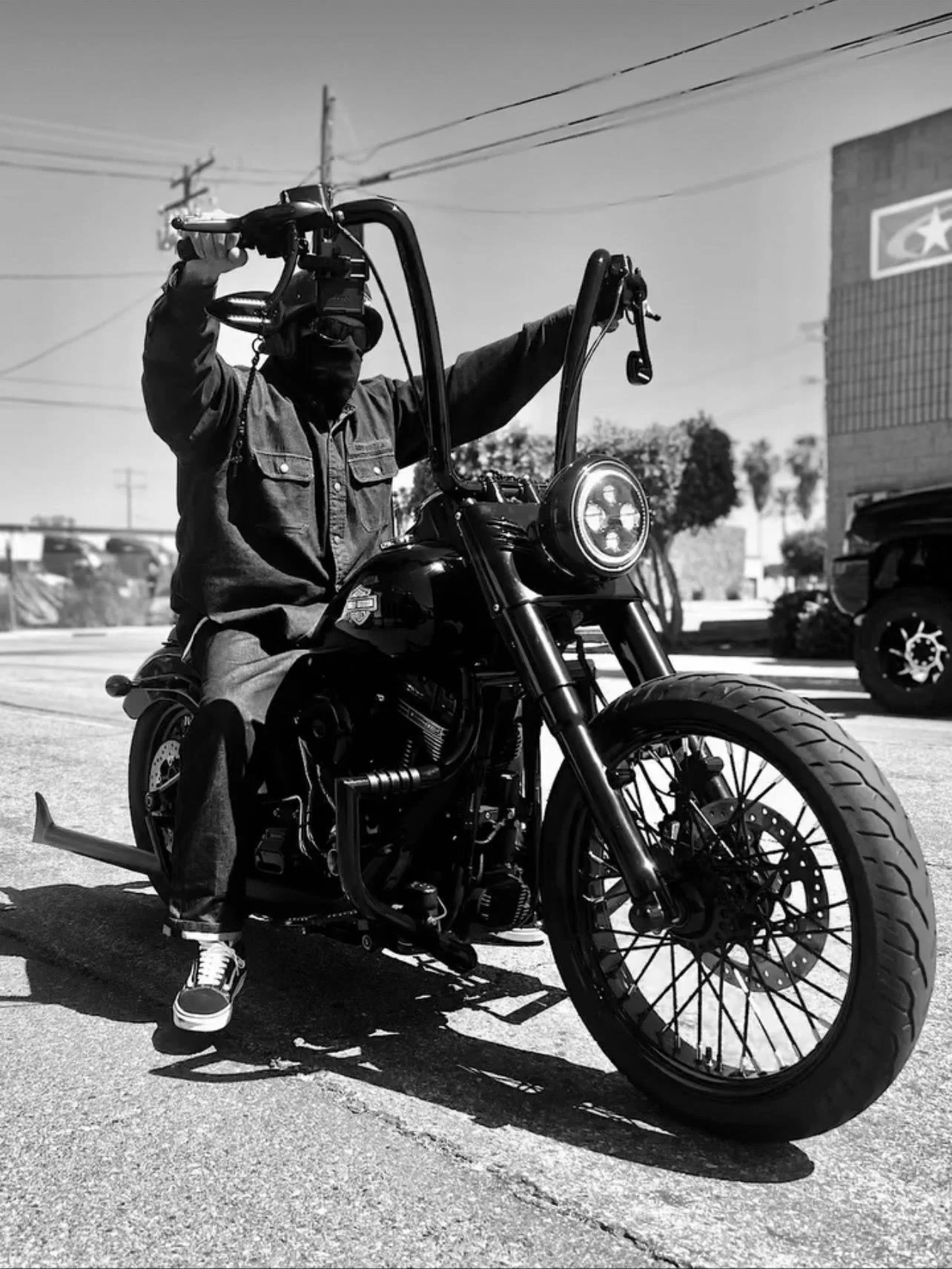 Person riding a black motorcycle on the street, wearing a dark jacket, face mask, and sneakers, with power lines and a building in the background.
