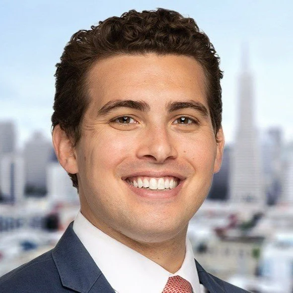 Professional man with dark curly hair and a bright smile, wearing a suit and tie, against a cityscape background.