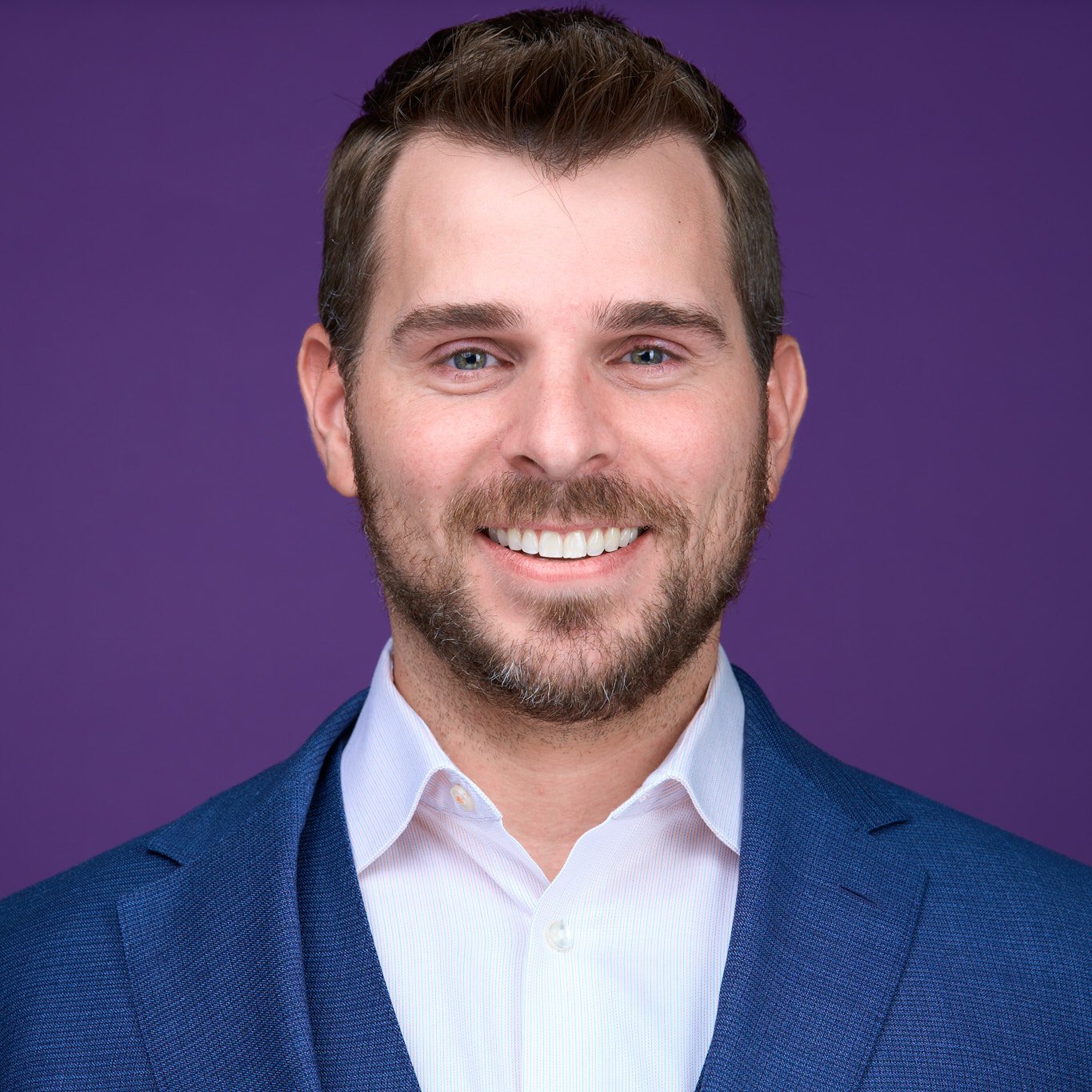 Formal portrait of a man with short brown hair and a beard, smiling, wearing a blue suit and white shirt, against a purple background.