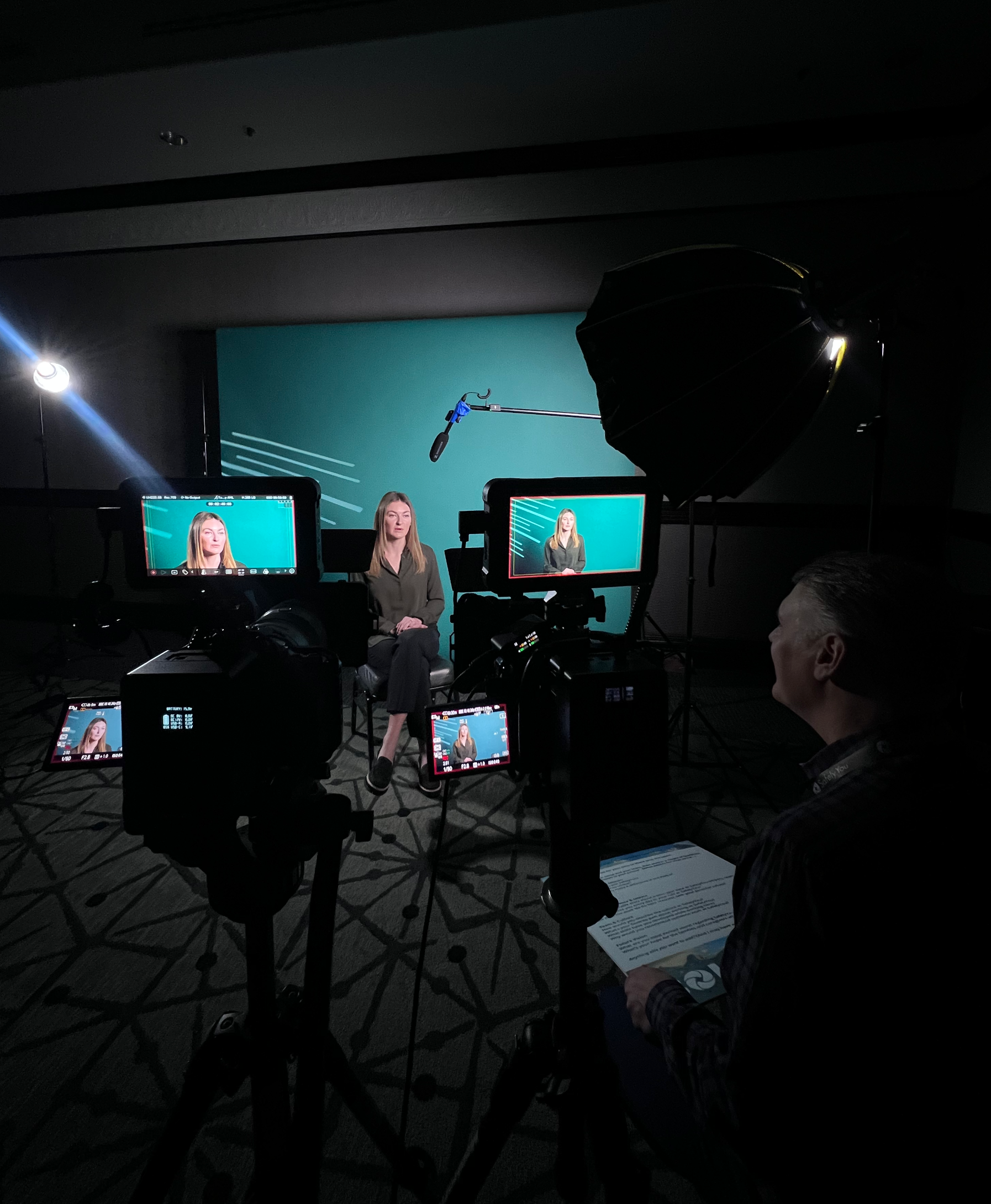A woman being filmed in a studio with three cameras, set against a green screen background. She is seated on a chair, facing the cameras, with filming equipment and a crew member visible.