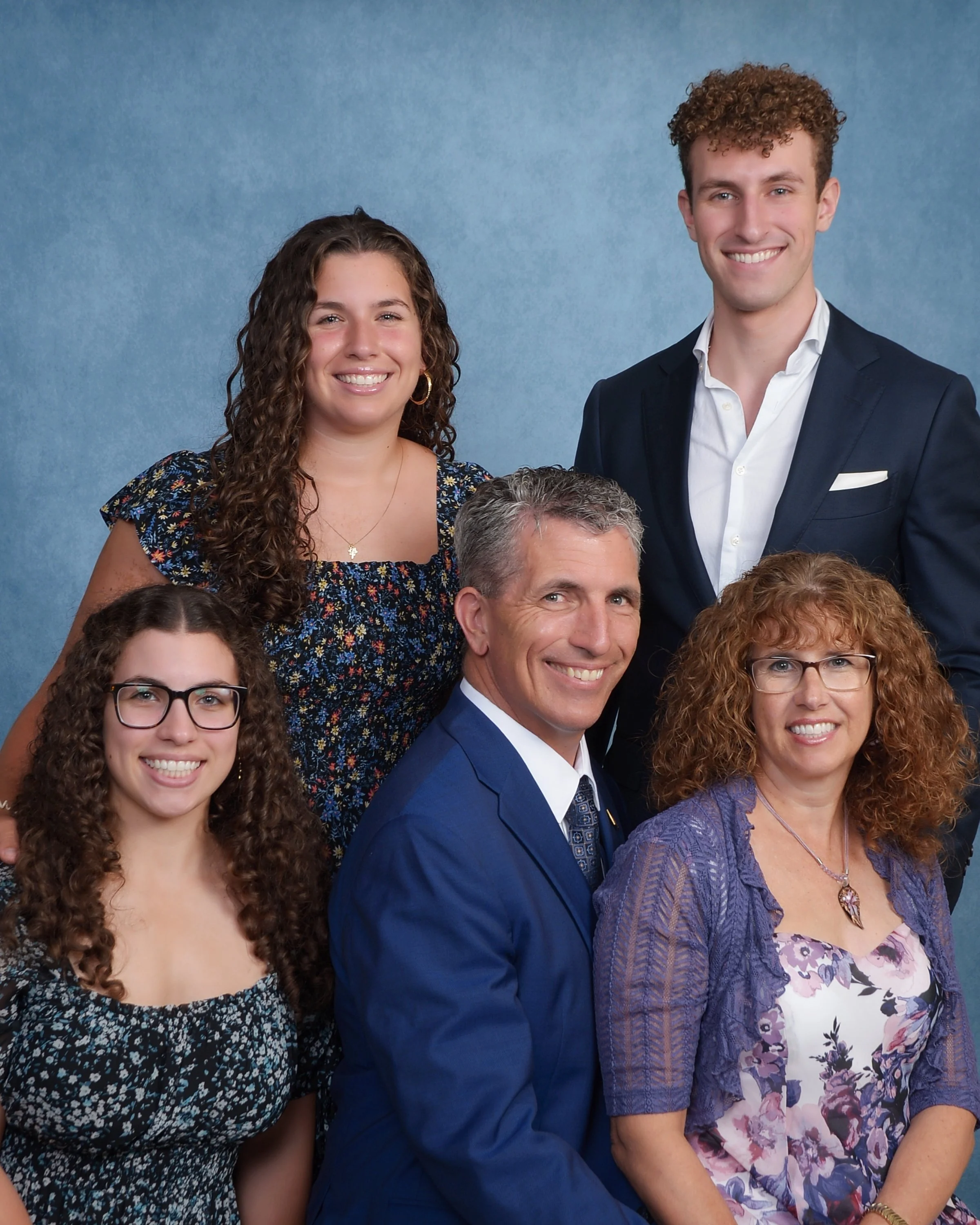 Group portrait of six people, three women and three men, dressed in formal attire, smiling against a blue background.