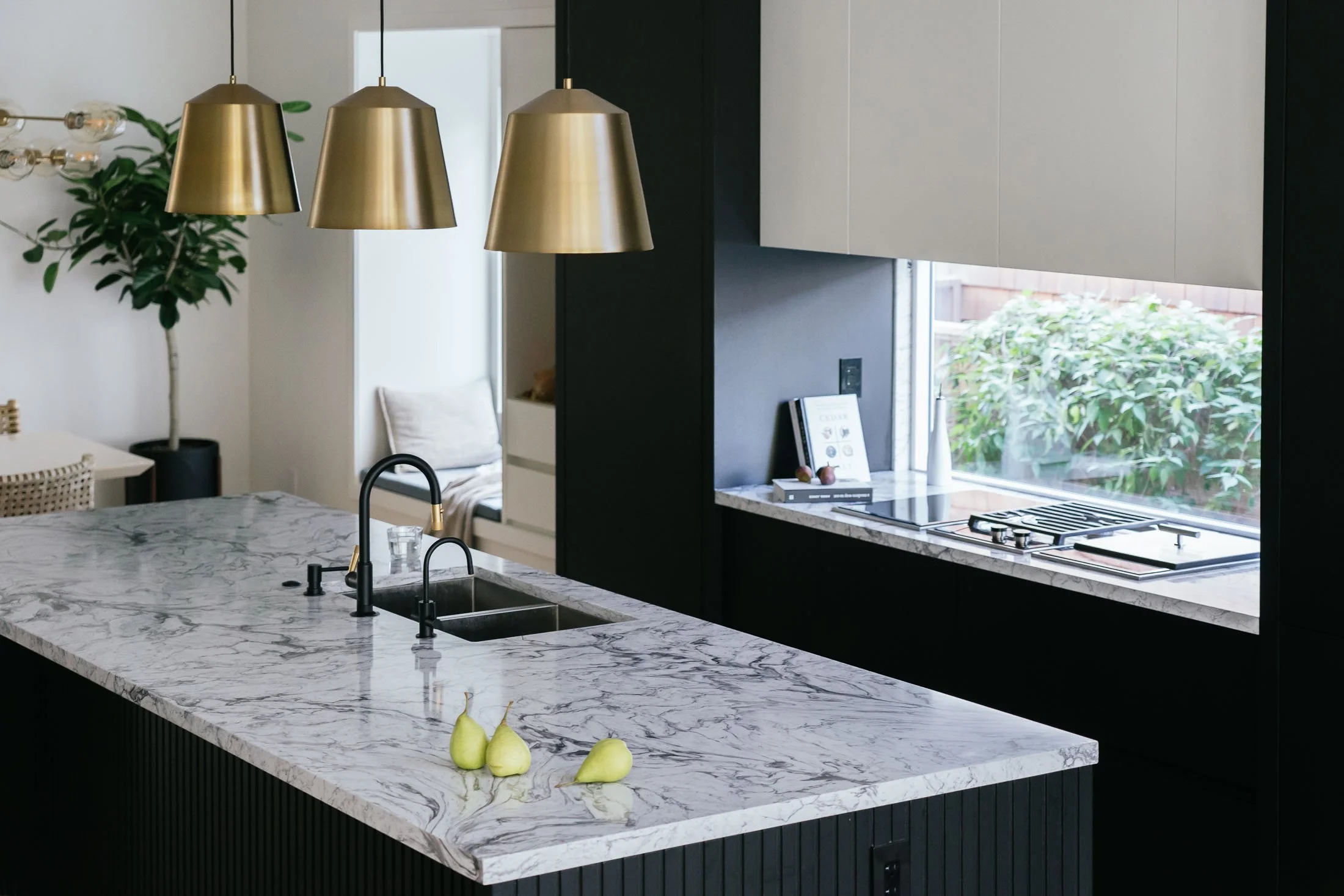 Modern kitchen with a marble island, black cabinets, and gold pendant lights. Green pears are on the island, and a window shows greenery outside.