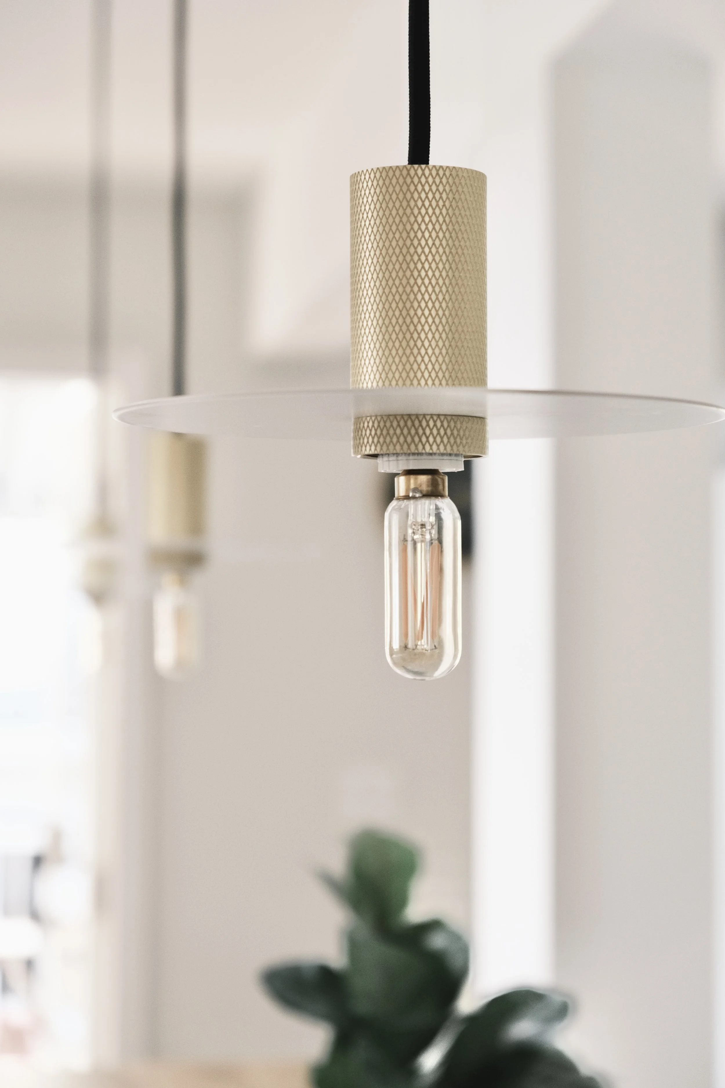 Close-up of a modern hanging light fixture with a cylindrical beige shade and a visible lightbulb, in a bright interior with a blurred plant at the bottom.