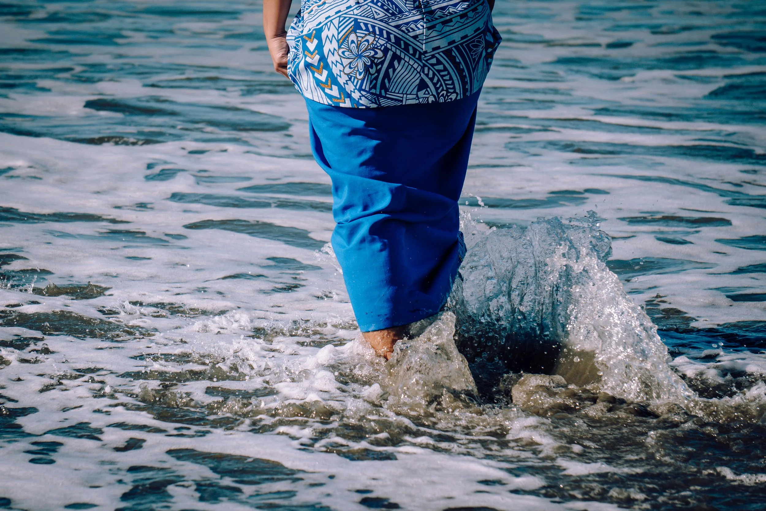 Person walking in the ocean with only their legs visible, wearing a blue patterned Puletasi in shallow water with small waves.