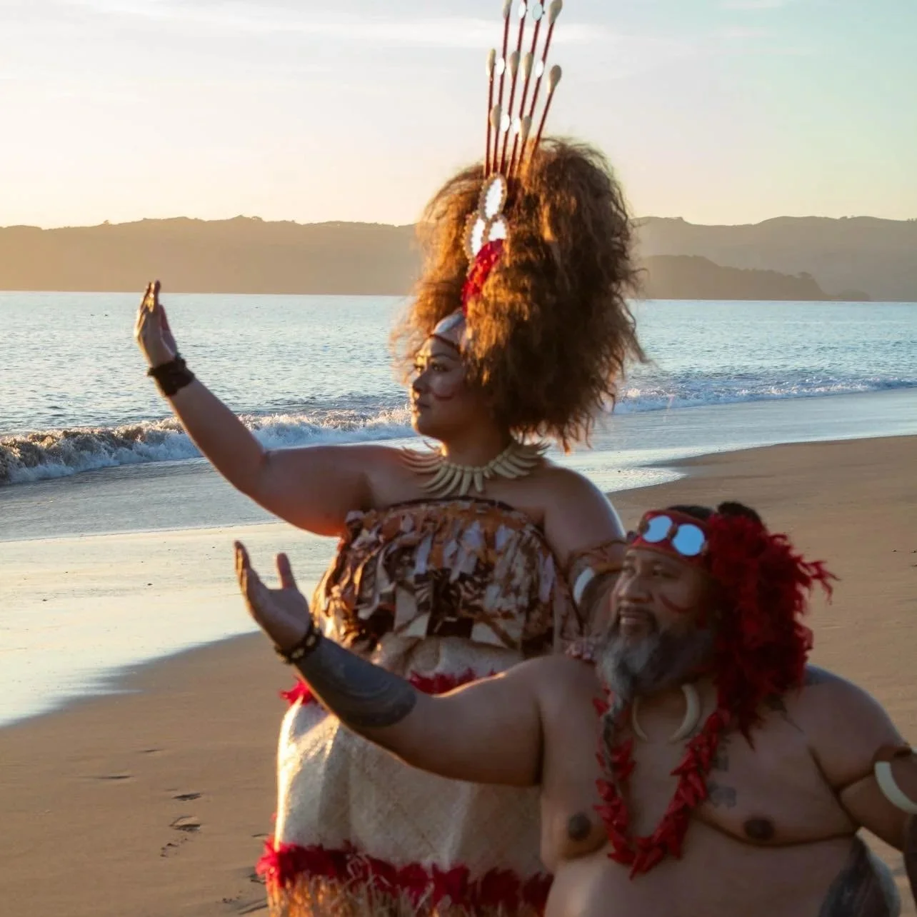Two Indigenous people, a man and a woman, dressed in traditional attire, performing a dance on the beach during sunset.