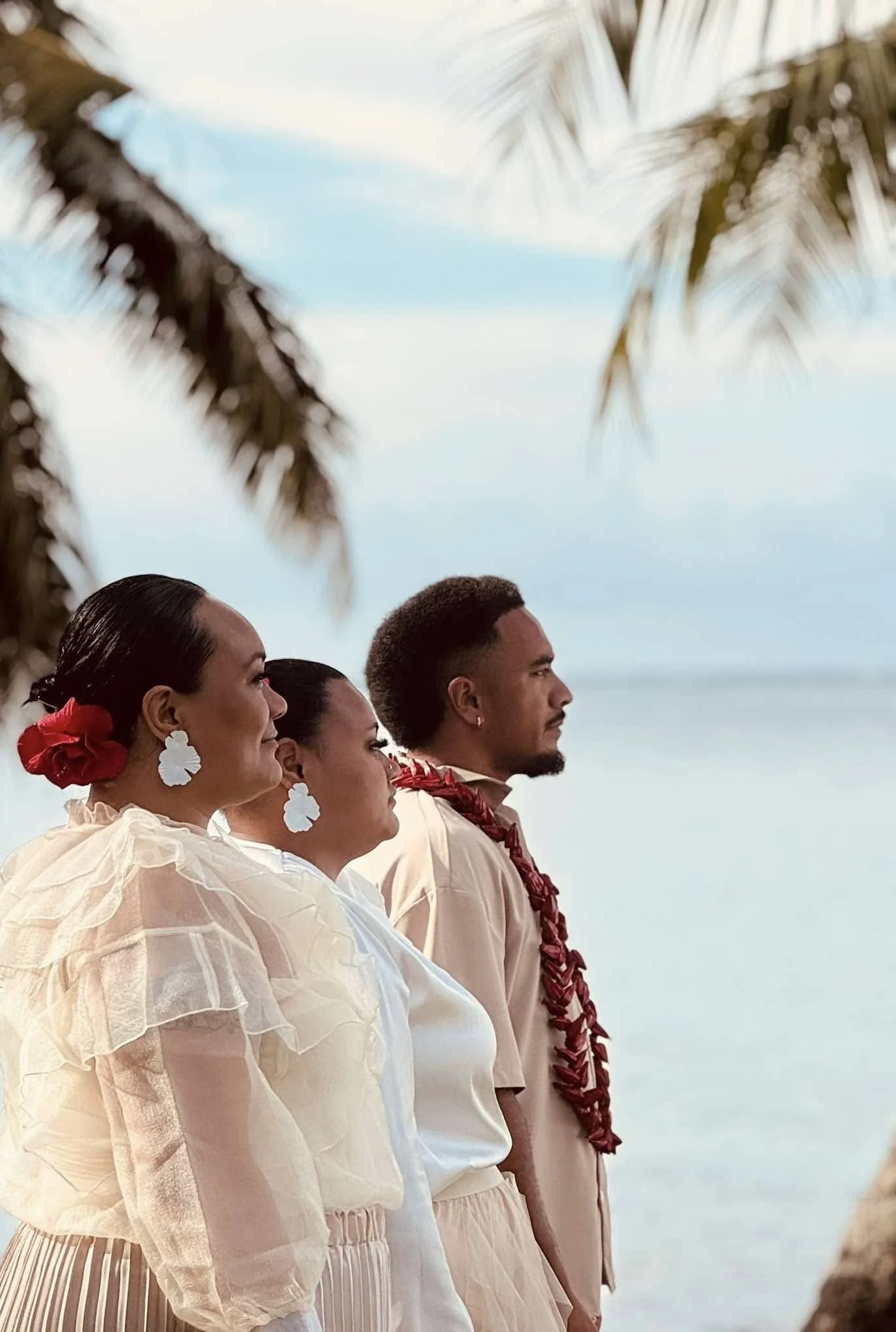 three people standing side by side outdoors, dressed in traditional clothing, near palm trees with a cloudy sky in the background.