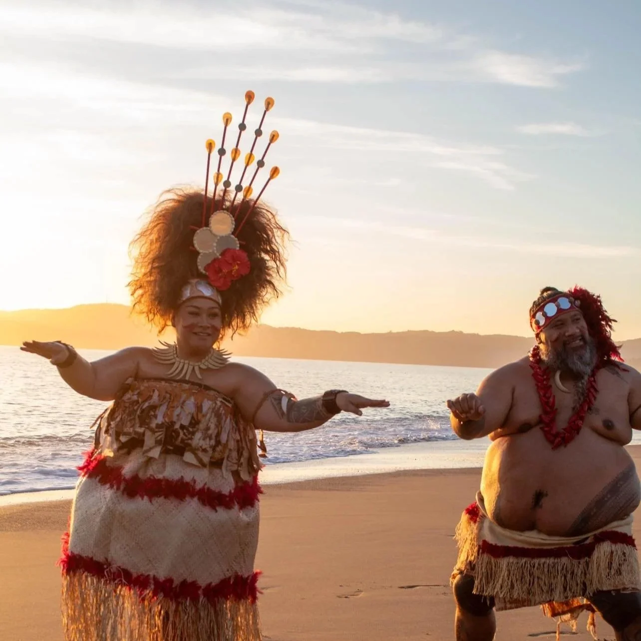 Two people dressed in traditional Polynesian attire dancing on a beach at sunset.