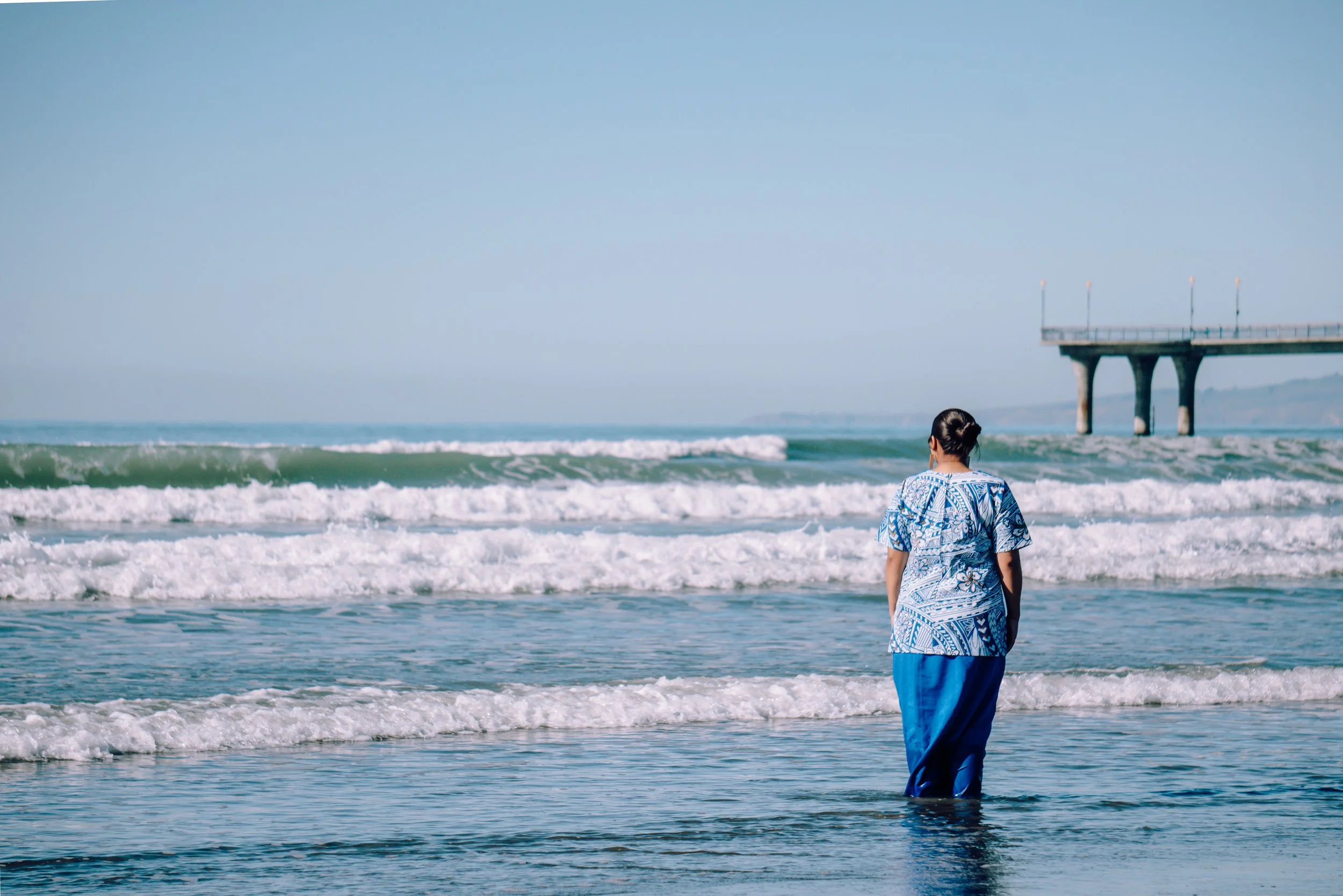 A woman in a blue patterned outfit walking on the beach towards the water, with waves in the background and a pier extending into the ocean.