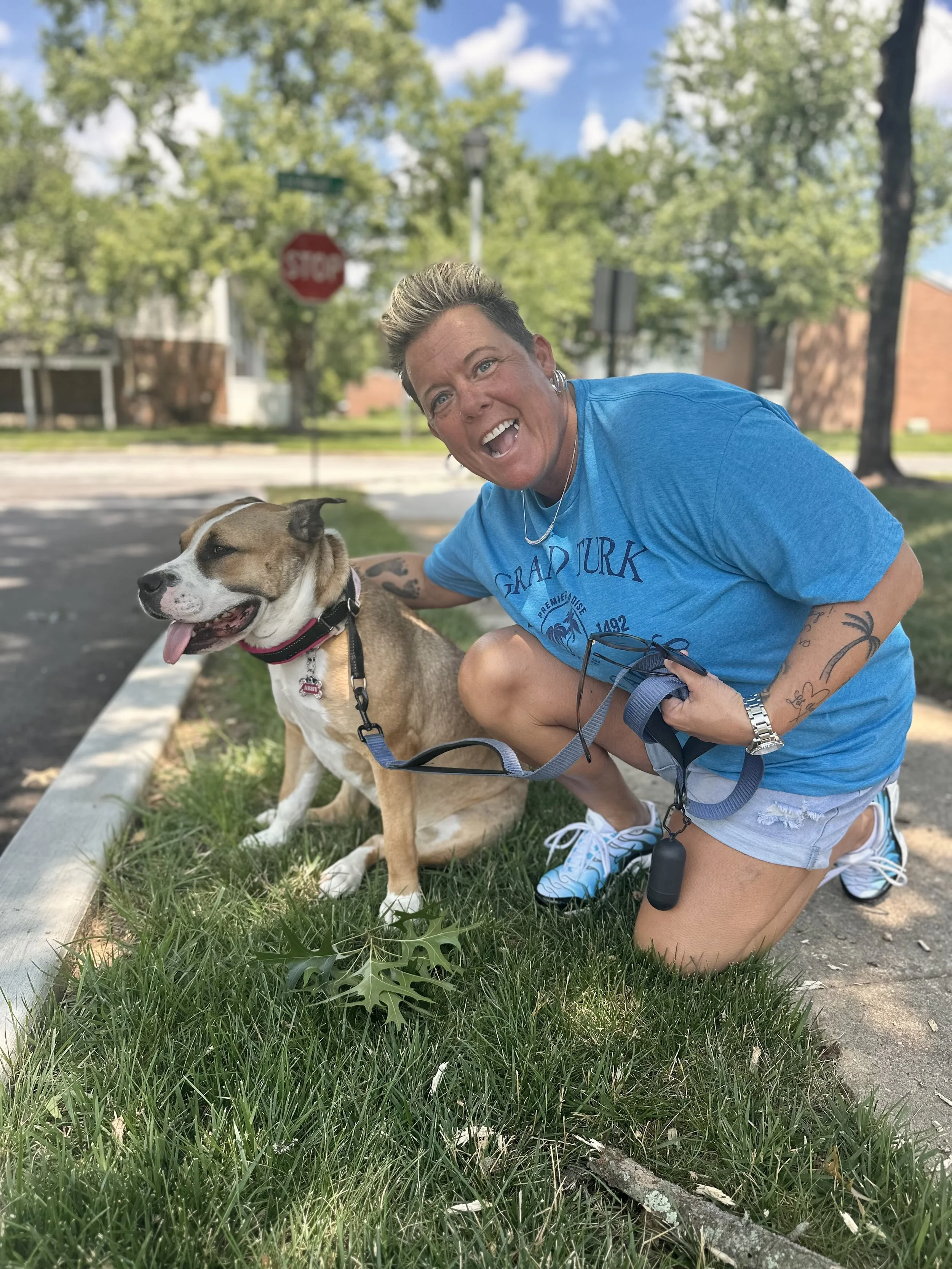 A woman with short blonde hair, wearing a blue T-shirt, shorts, and sneakers, squats next to a smiling dog with a tan and white coat, on a leash, outdoors on a grassy area with trees and a street in the background.