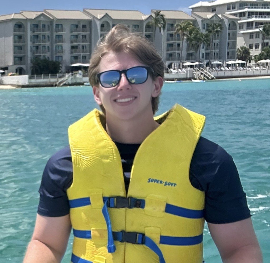 A young man with sunglasses smiling while wearing a yellow life jacket on a boat, with a marina, palm trees, and resort buildings in the background on a sunny day.