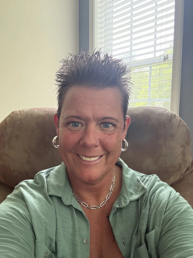 A woman with short spiky hair, blue eyes, and silver hoop earrings, smiling while sitting on a beige armchair near a window with blinds.