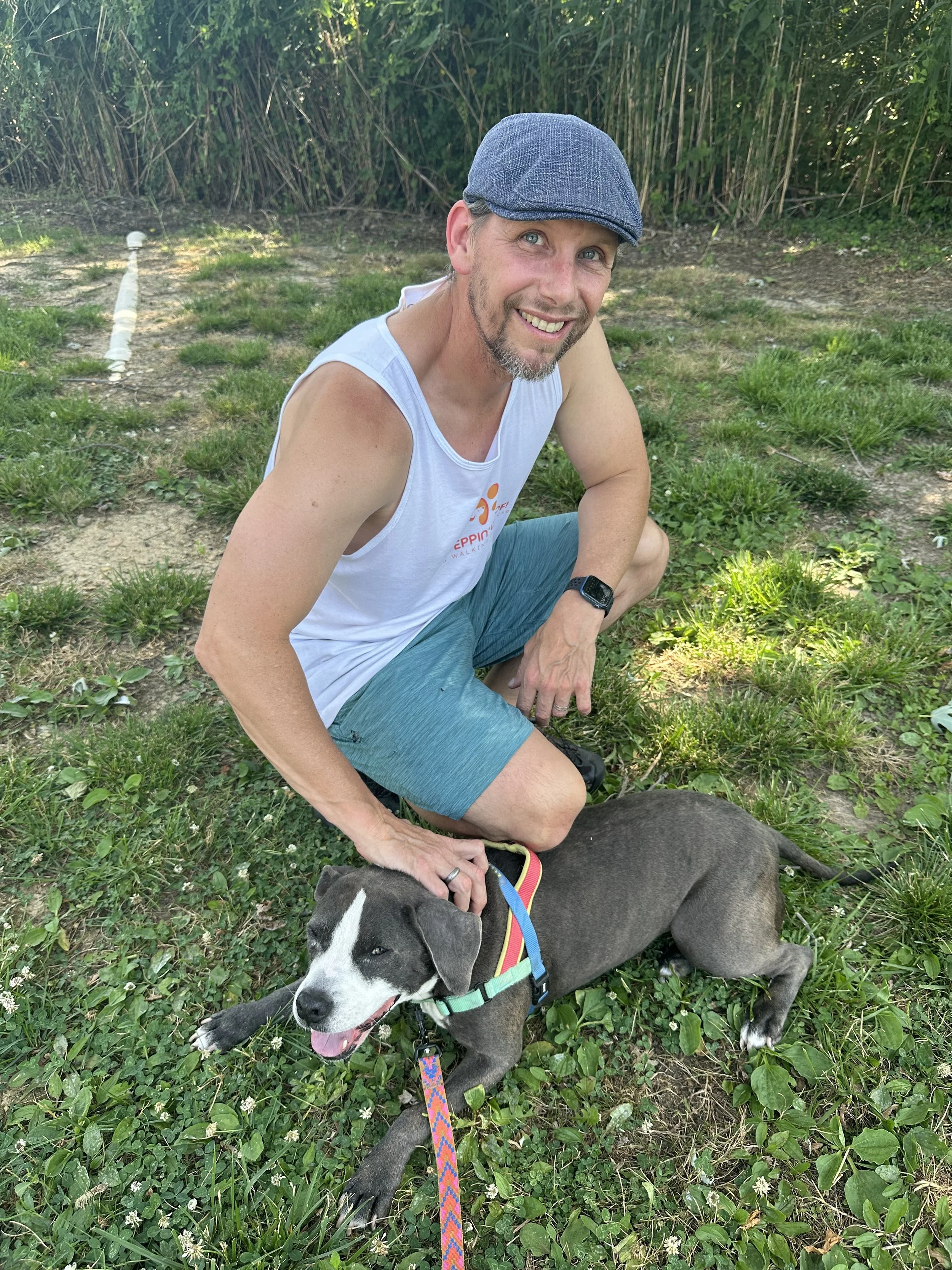 A man with light skin, wearing a gray flat cap, sleeveless white shirt, blue shorts, and a black watch, crouching outdoors on grass with lush green bushes in the background. He is smiling and petting a gray and white puppy lying on the grass, which is wearing a colorful collar and leash.