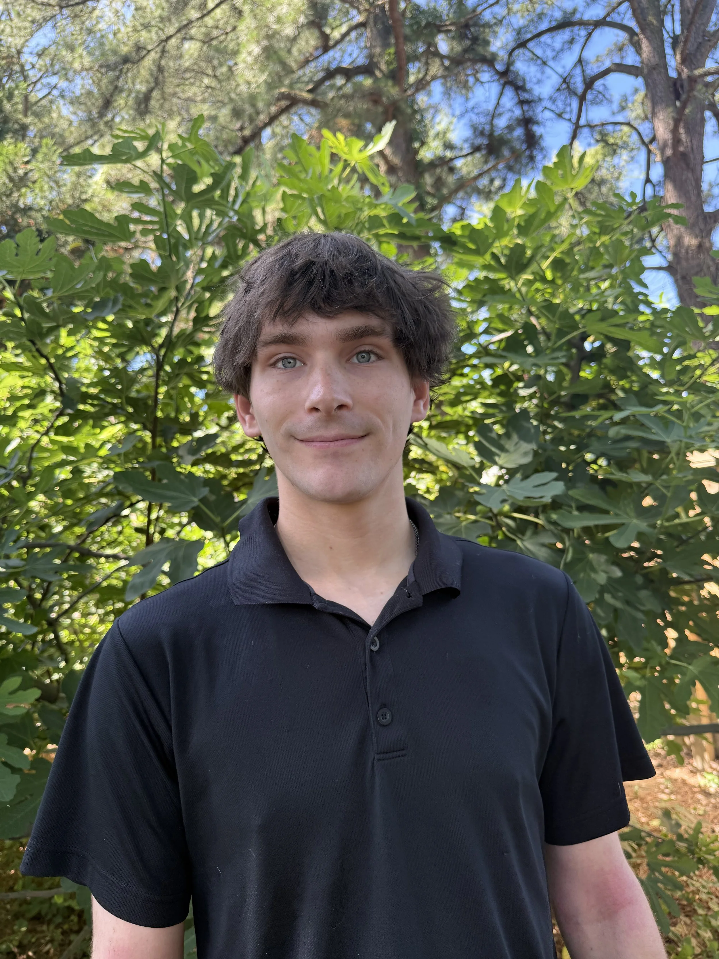 A young man with light skin and blue eyes standing outdoors among lush green foliage and trees, wearing a black collared shirt.