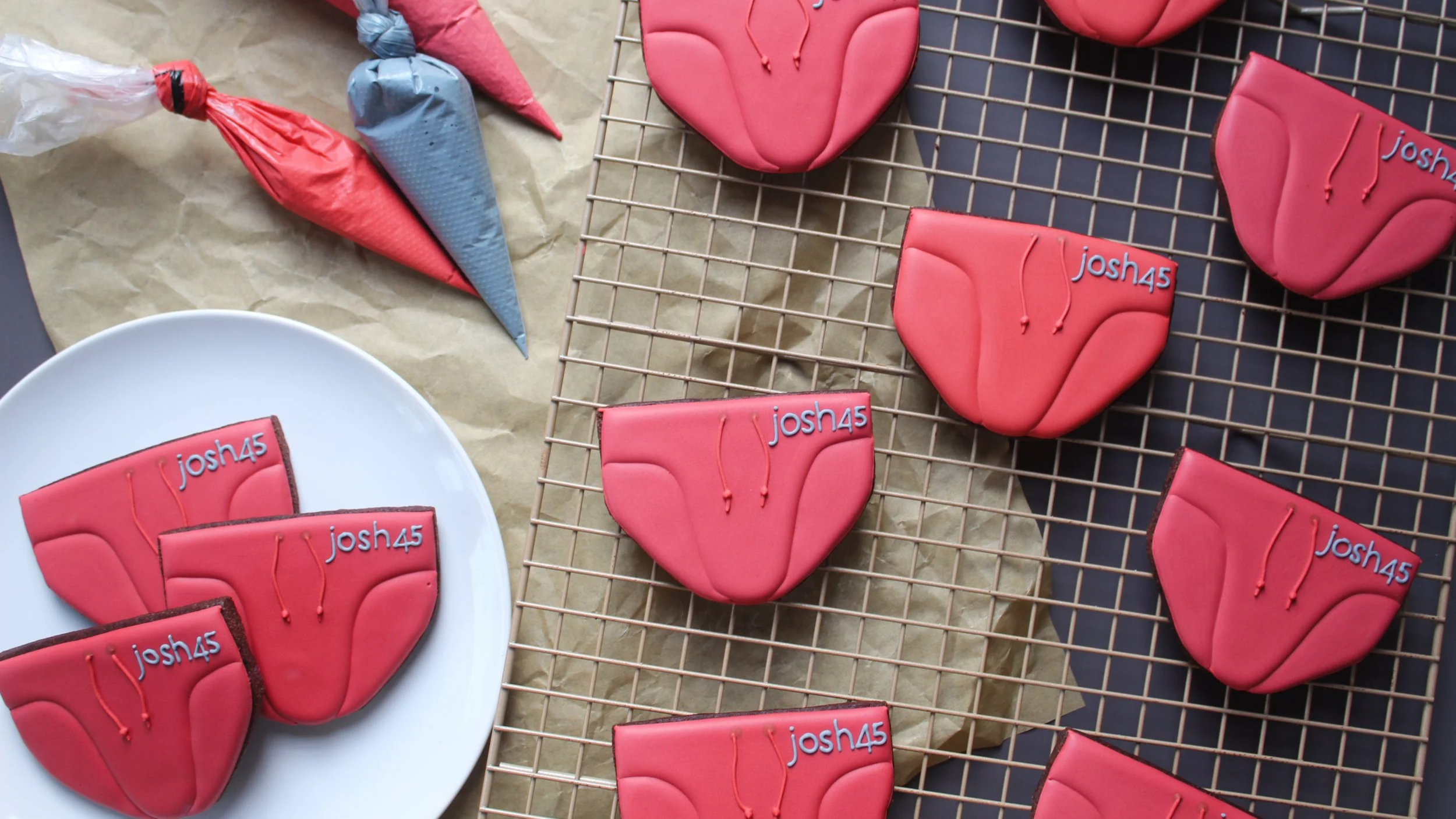 Decorative cookies shaped like speedos with red icing, each with the name 'josh45' written on them, placed on a cooling rack and a white plate, with cake decorating cones in pink and blue icing nearby.