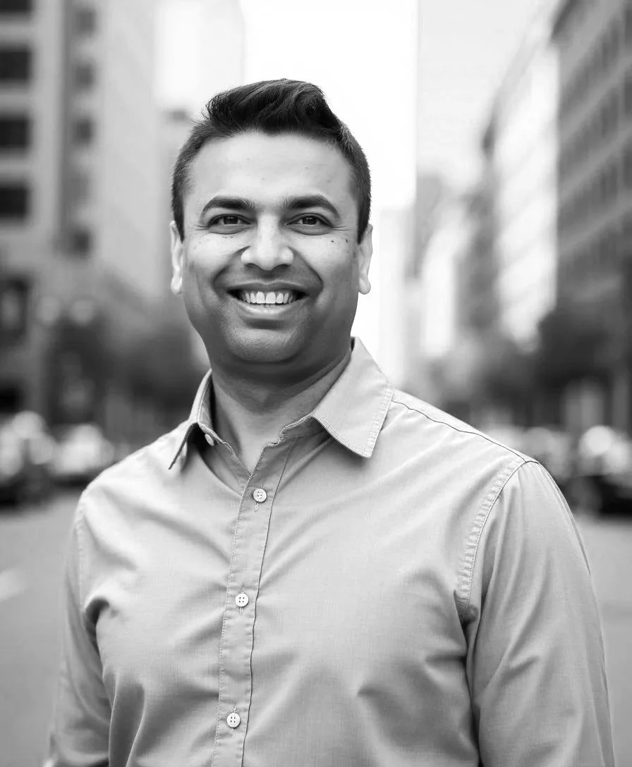 A smiling man in glasses with arms crossed, standing in a home office with a wooden desk, computer monitors, and a window in the background.