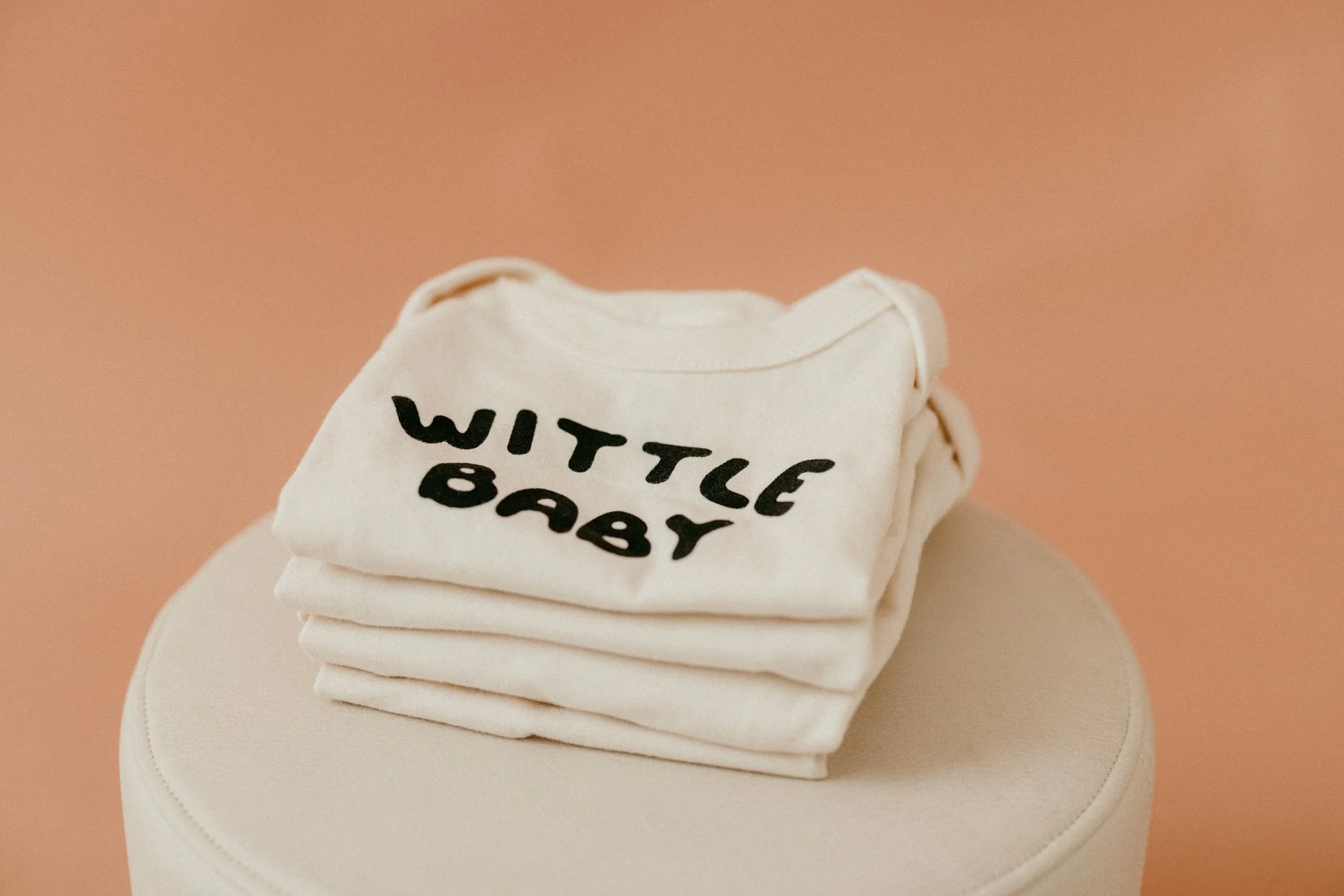 Stack of white t-shirts with black text that says 'WITTLE BABY' on a round cream-colored stool against a soft pink background.