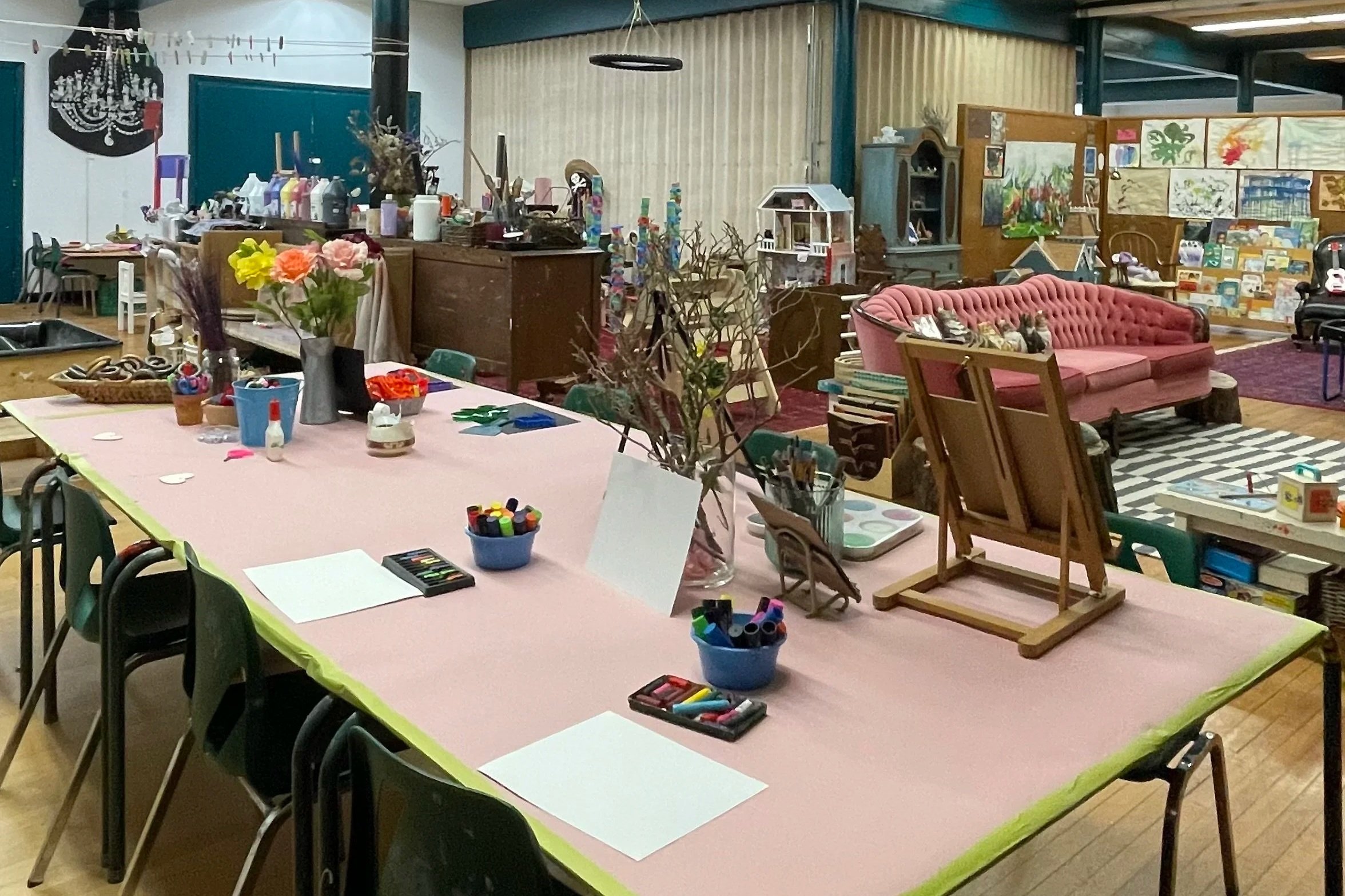 A large table covered with pink craft paper, surrounded by chairs, in a room filled with arts and crafts supplies, decorations, and furniture including a pink sofa and art displays.