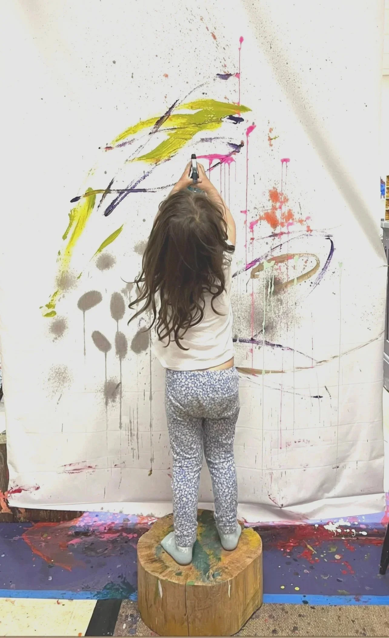 A young girl standing on a wooden block, painting an abstract artwork on a large white canvas with various colors.