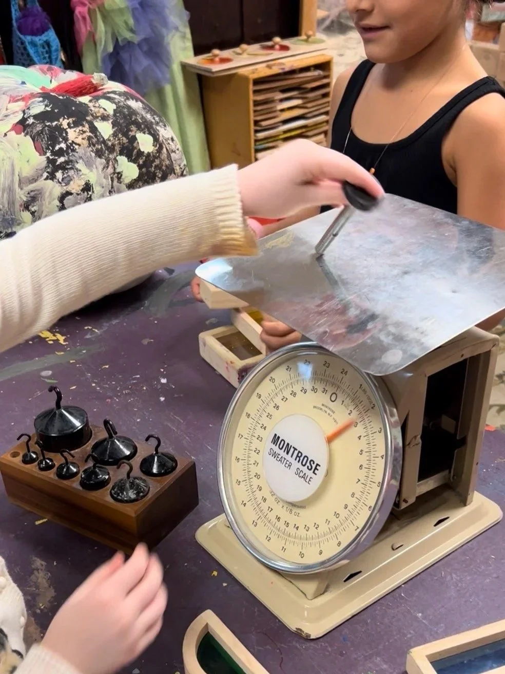A person is weighing an object on a vintage scale labeled 'Montrose Sweater Scale.' A child in a black tank top looks on as another person holds a metal sheet, possibly being weighed or examined. Various small black objects are on a wooden block nearby, and the background shows colorful clothing and shelves.